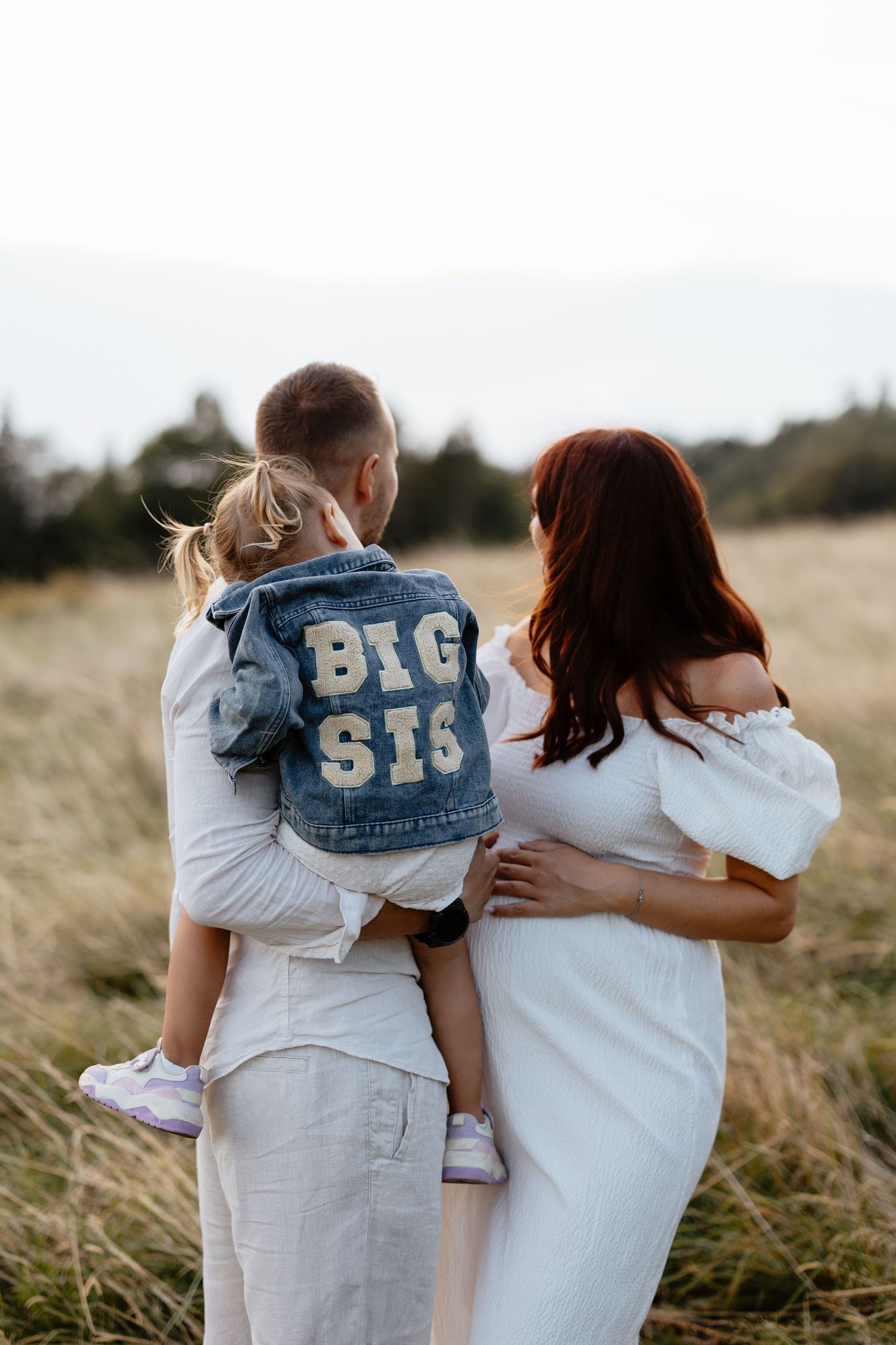 A family in white outfits poses together, with child wearing a 'Big Sis' denim jacket.