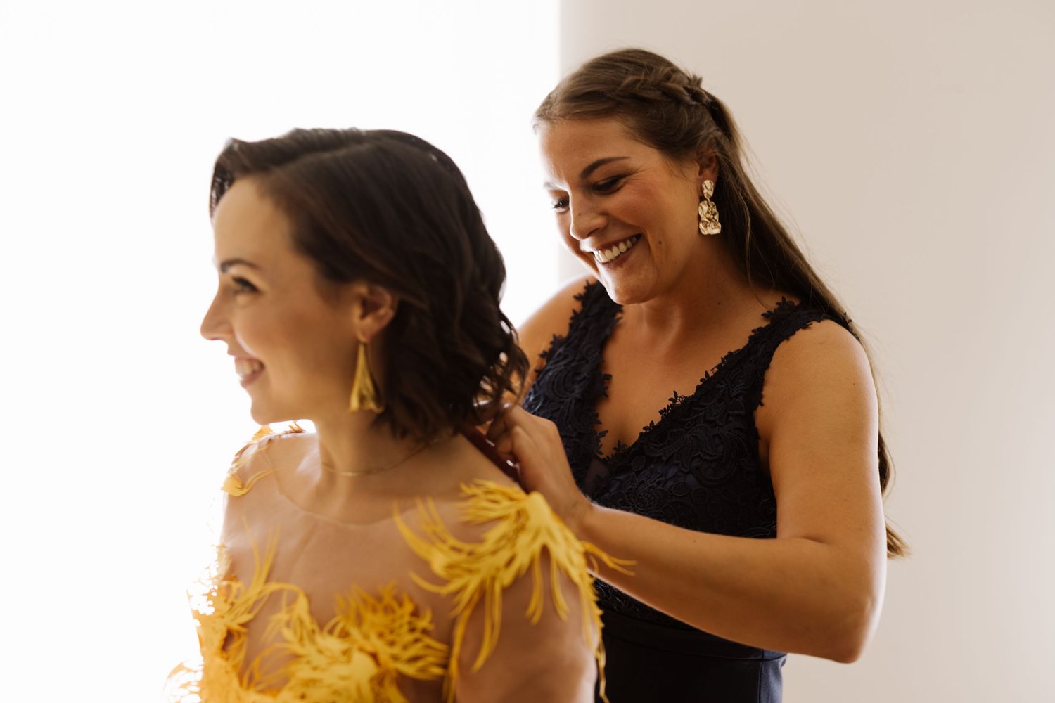 A woman in a navy lace dress helps another woman in a yellow floral dress get ready for a special occasion.