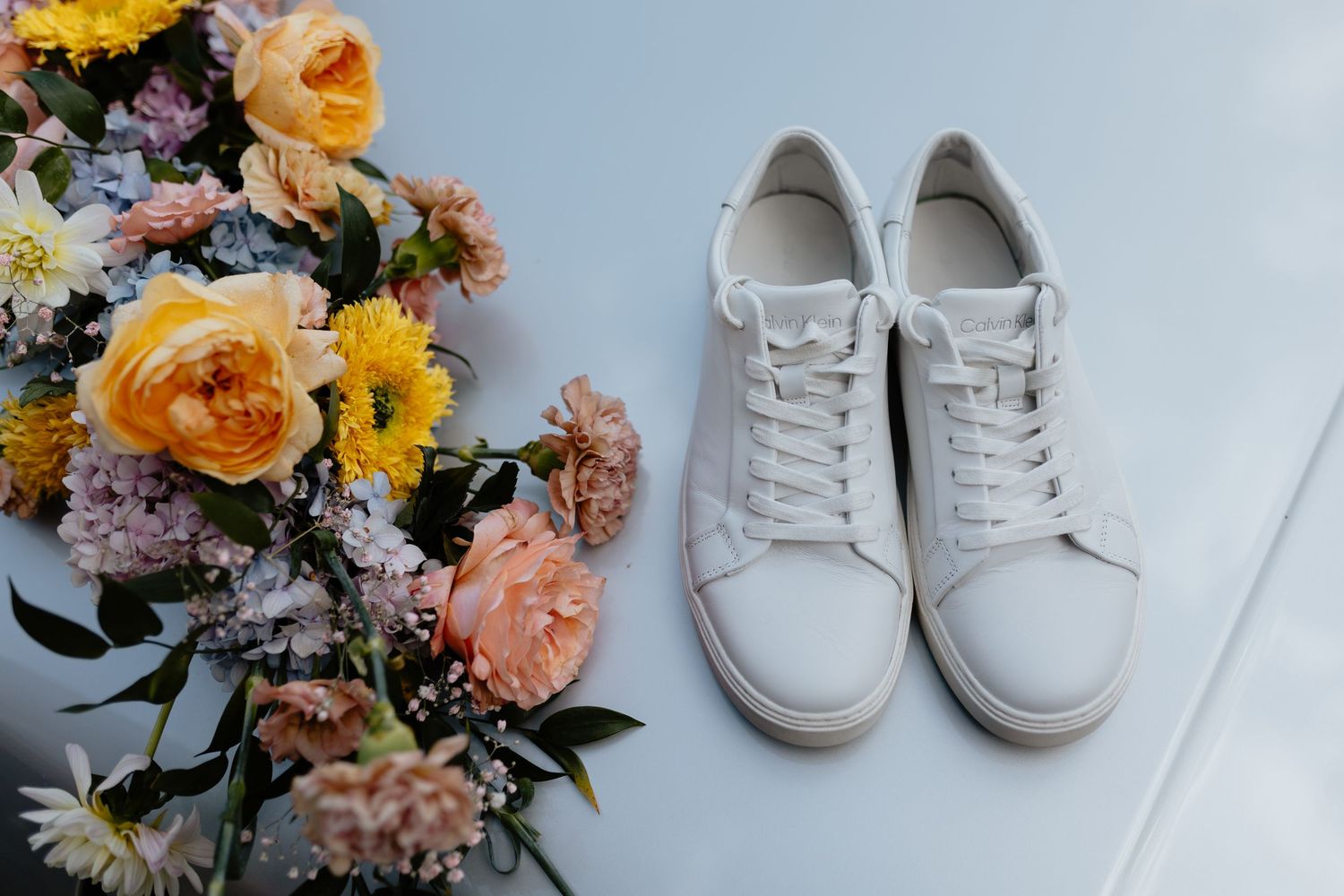 A pair of white baby shoes surrounded by colorful floral arrangements with yellow, peach and pink blooms.