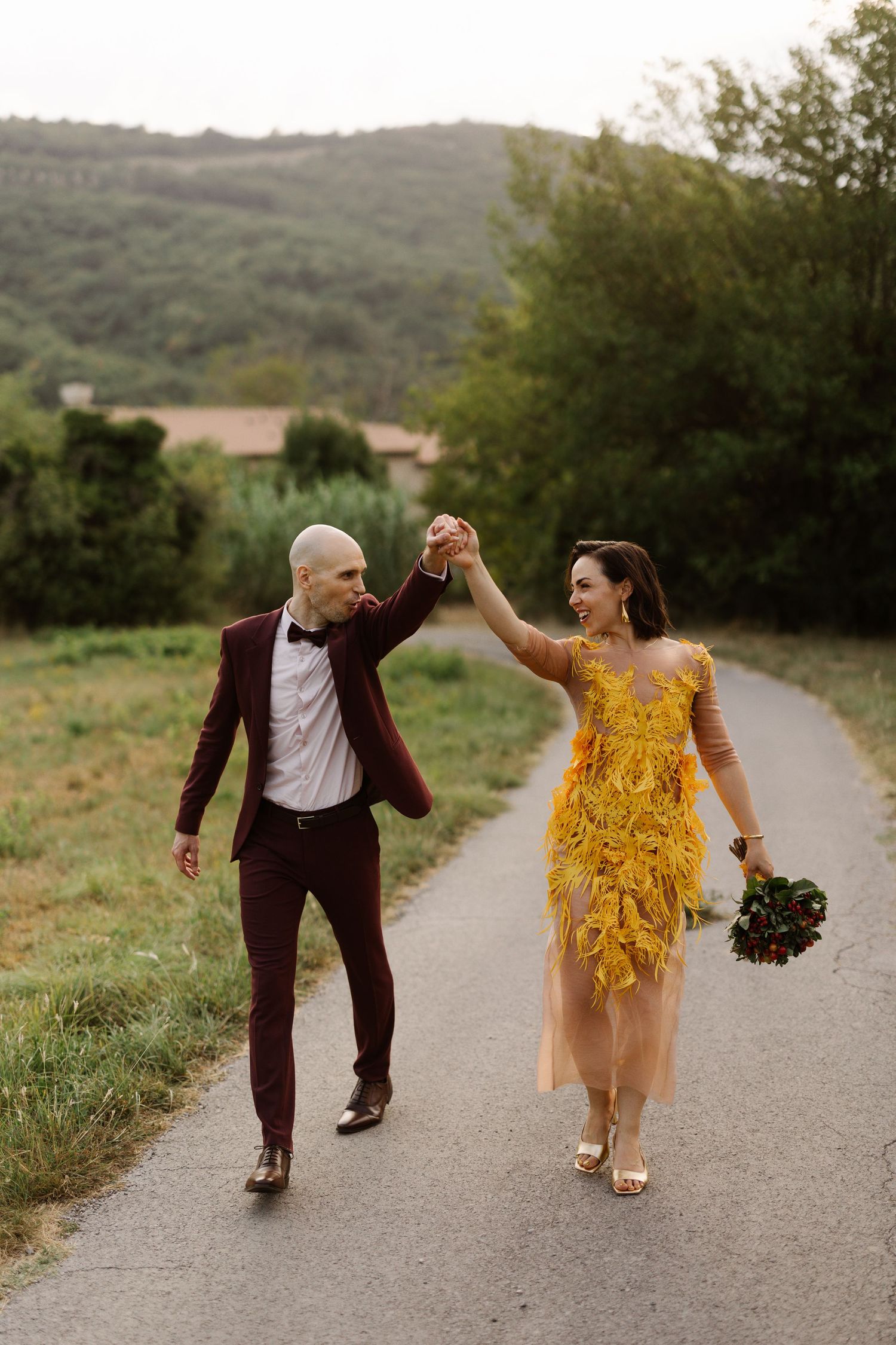 Couple walking and dancing on path wearing burgundy suit and yellow dress.