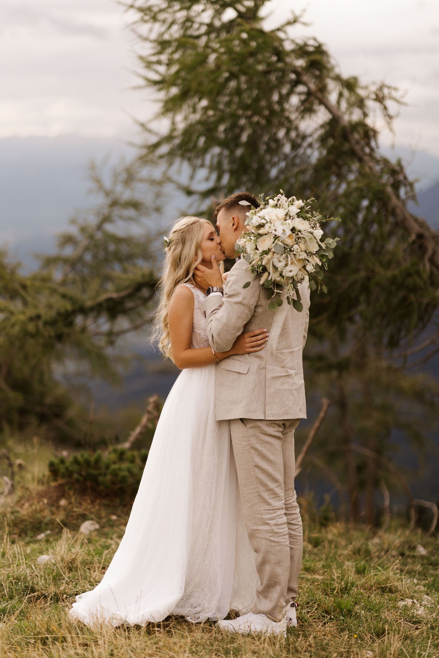 A romantic wedding couple embraces on a hilltop with mountain views in the background.