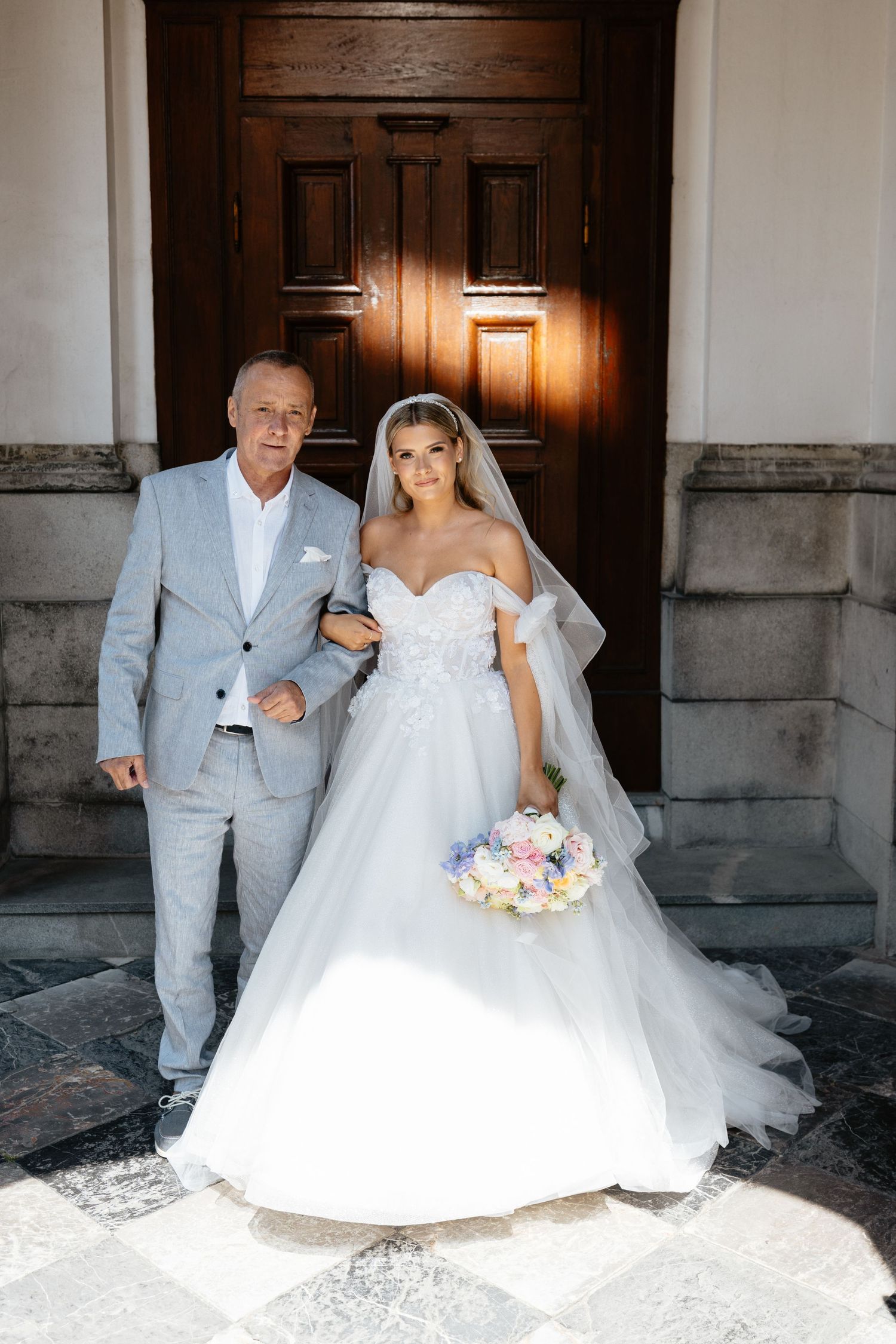 A bride in a white ballgown and groom in light gray suit pose together in an elegant stone archway.