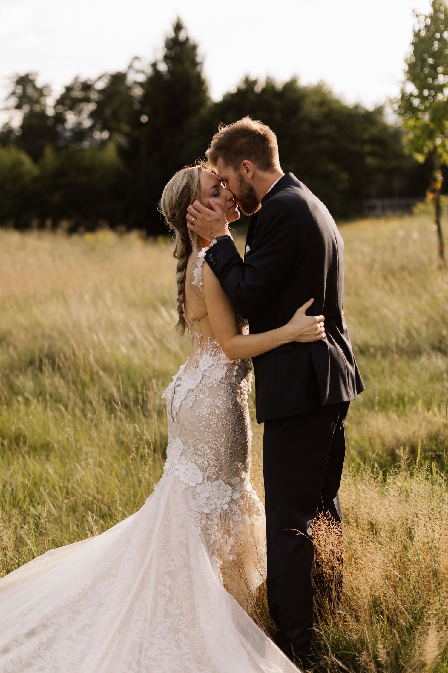 A romantic wedding couple embraces in a sunlit meadow with tall grass and trees in the background.