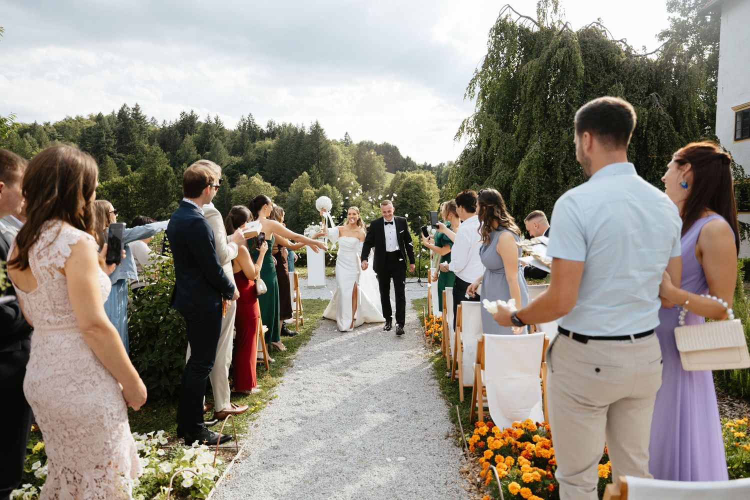 Wedding guests line a path outdoors with colorful flowers and decorations during a summer ceremony.