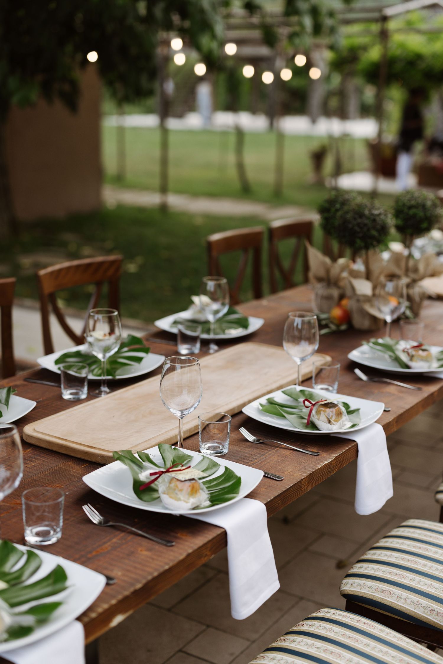 An elegant outdoor wedding table setting with string lights, green leaves and wooden chairs.
