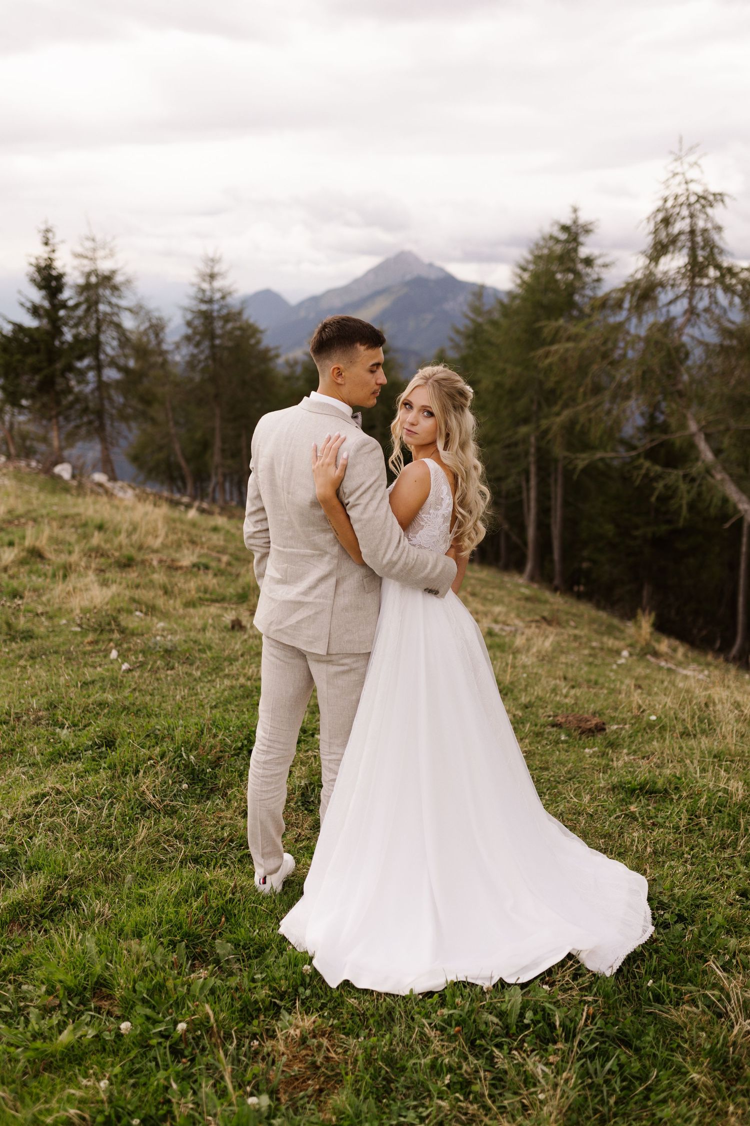 A romantic wedding moment captured on a scenic mountain hillside with evergreen trees in the background.