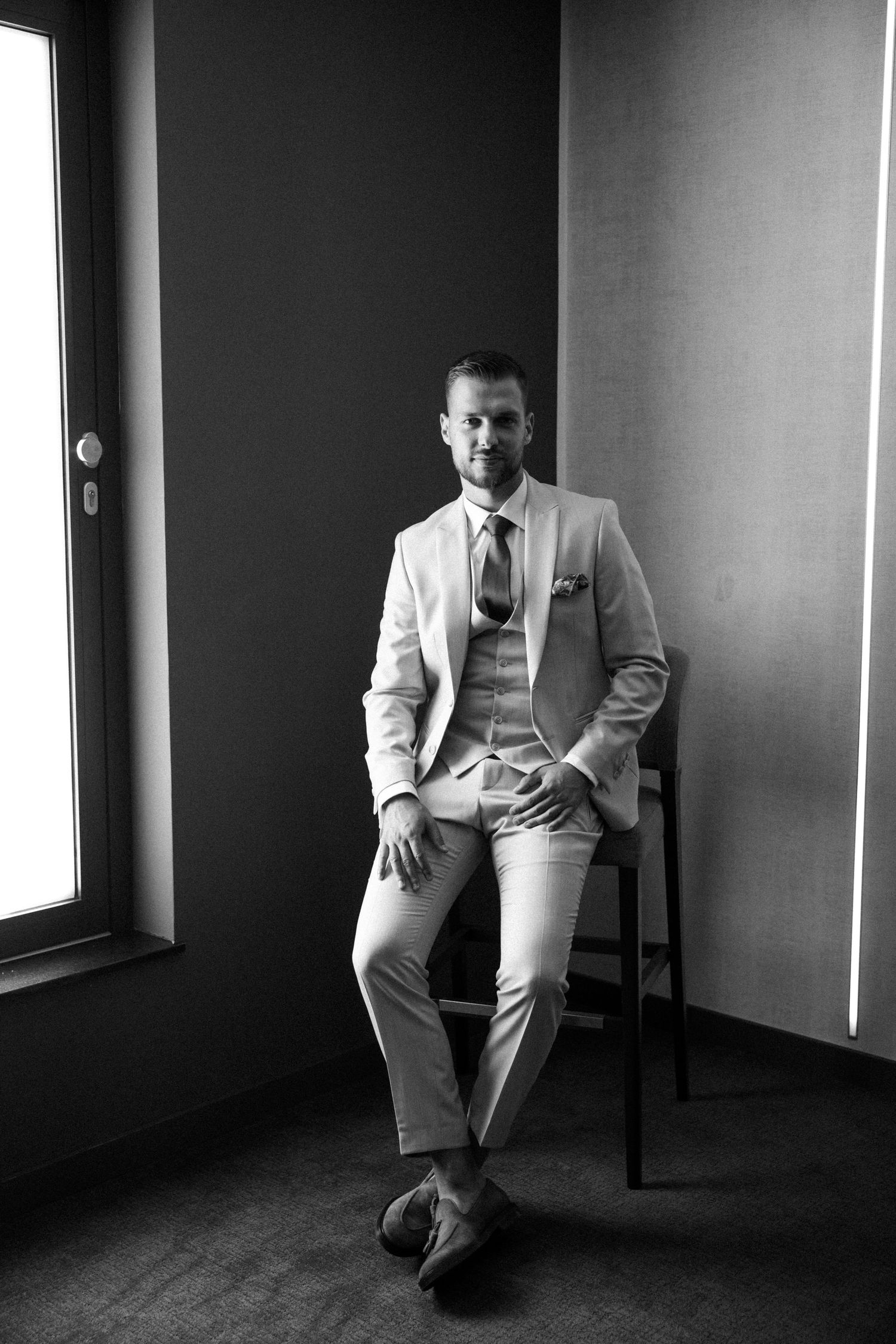 A groom in a light beige suit sits on a chair in a minimalist setting for a black and white portrait.