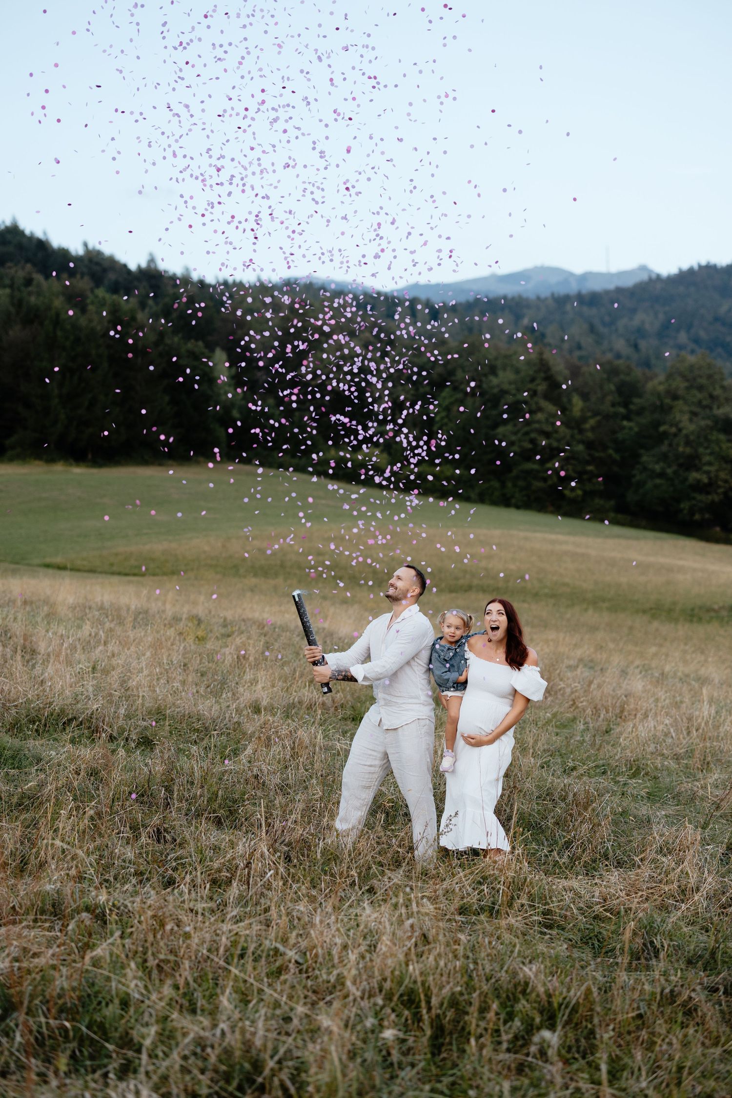 A couple in white outfits celebrates with confetti in a scenic mountain field.