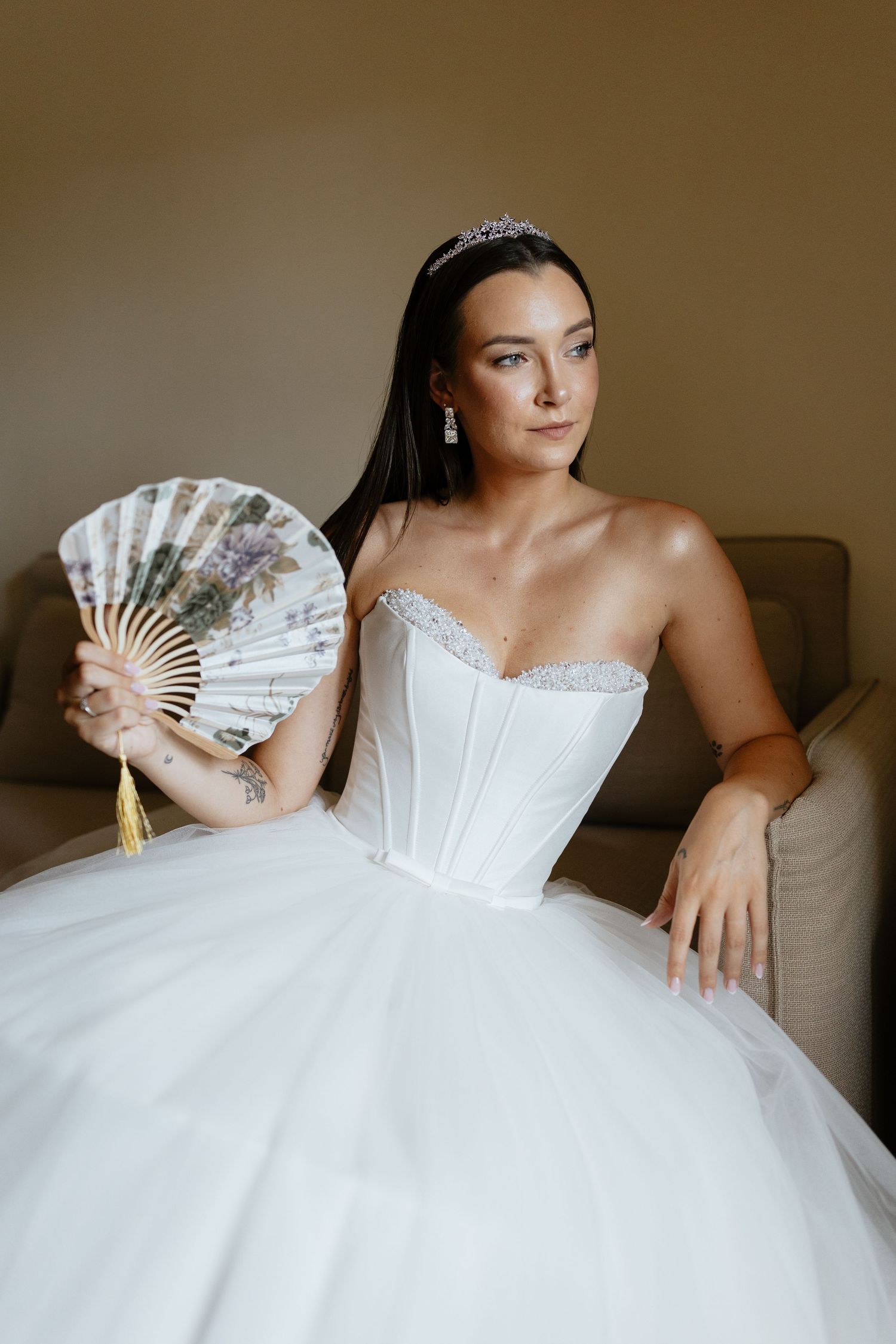 Bride in a white strapless ball gown poses with a decorative fan on a couch.