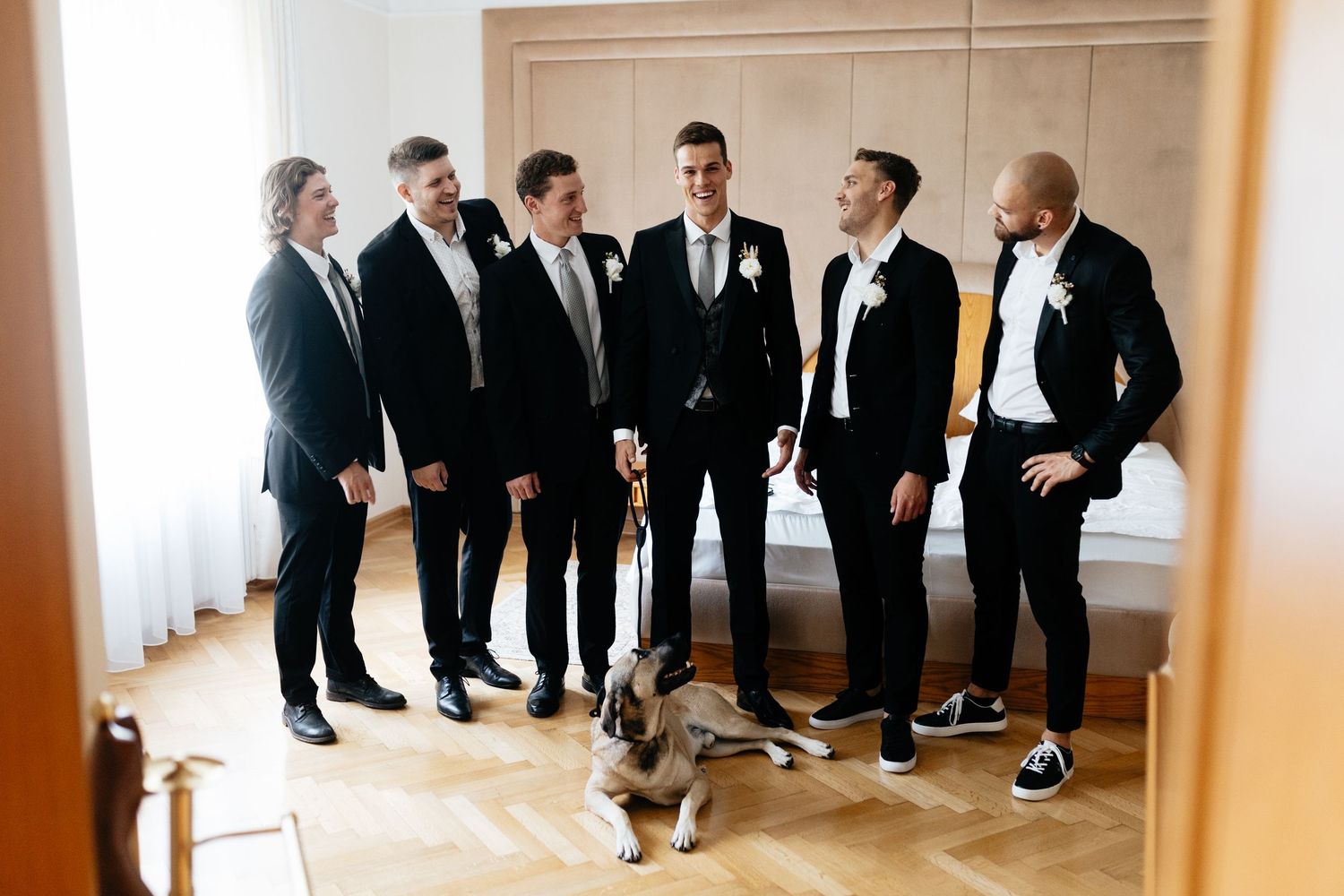 Groomsmen in black suits and dress shoes gather in a bright room for a candid wedding photo moment.