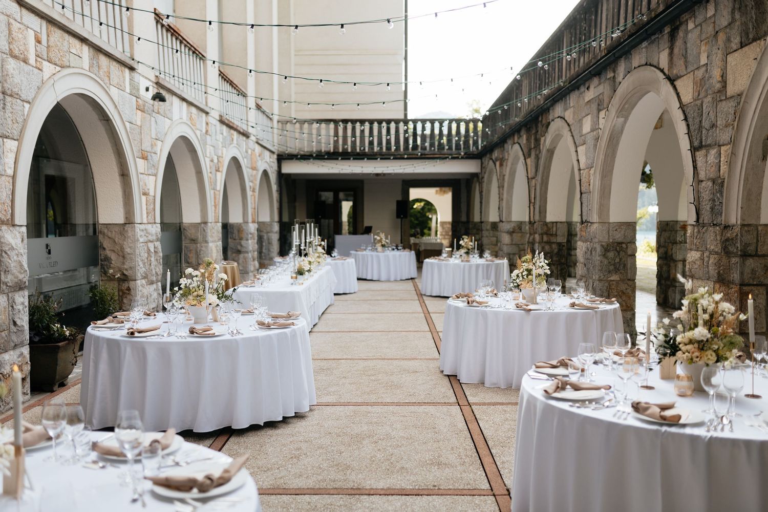 Outdoor wedding reception setup with white-clothed round tables and string lights in a stone courtyard.