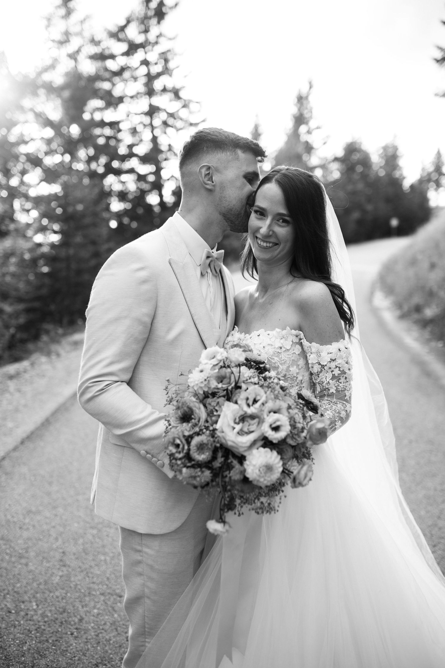 A wedding couple poses together outdoors with a beautiful floral bouquet against an evergreen backdrop.