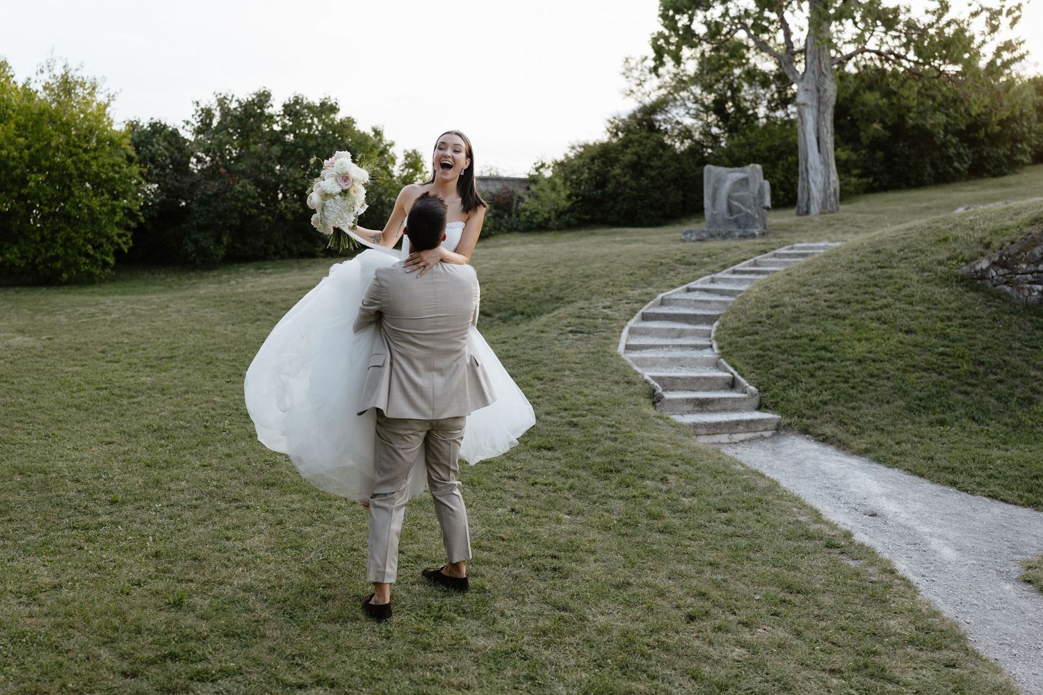 Newlyweds walk hand in hand down winding stone path surrounded by green landscape.