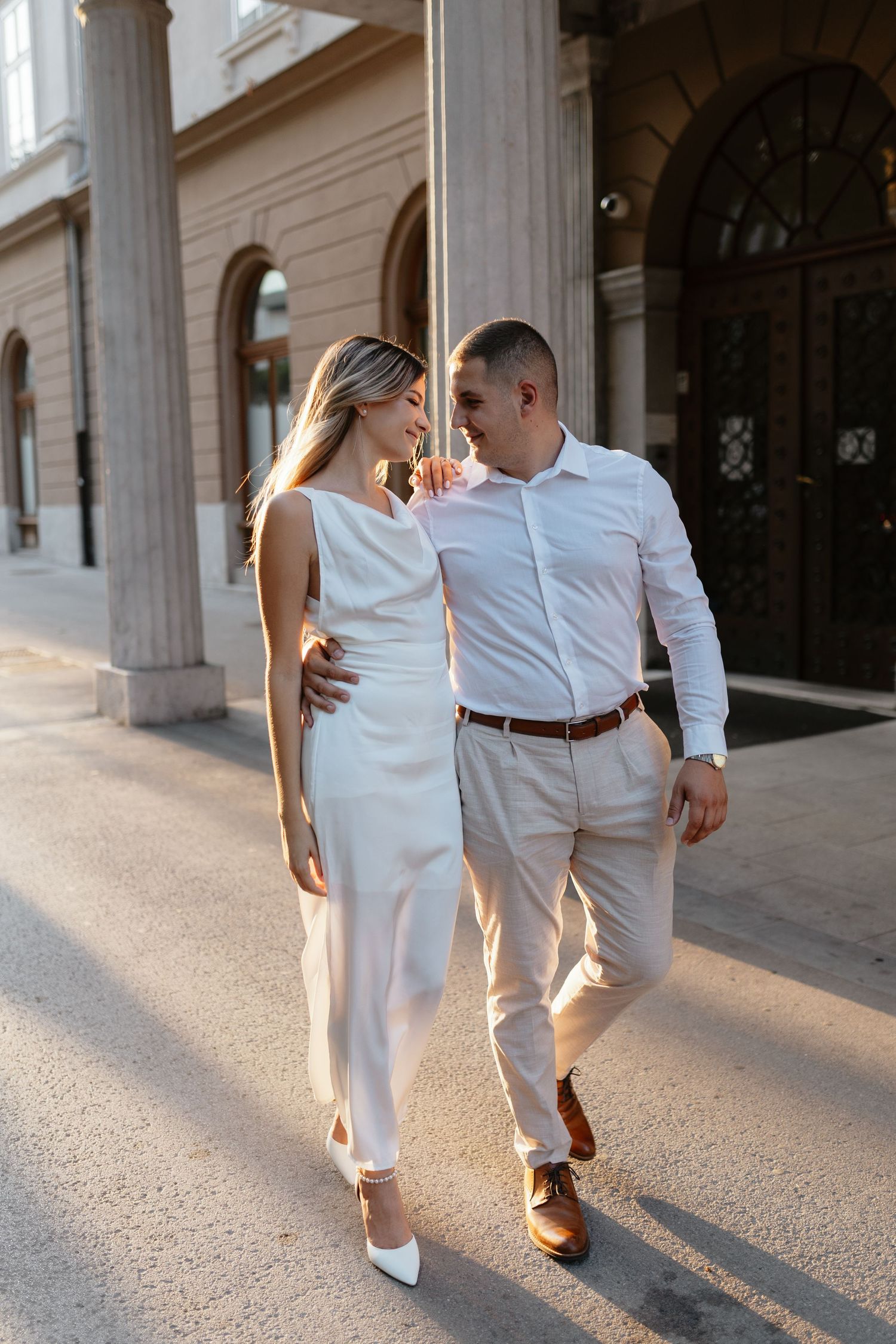 A couple in coordinated white outfits walking together along a classical building exterior.