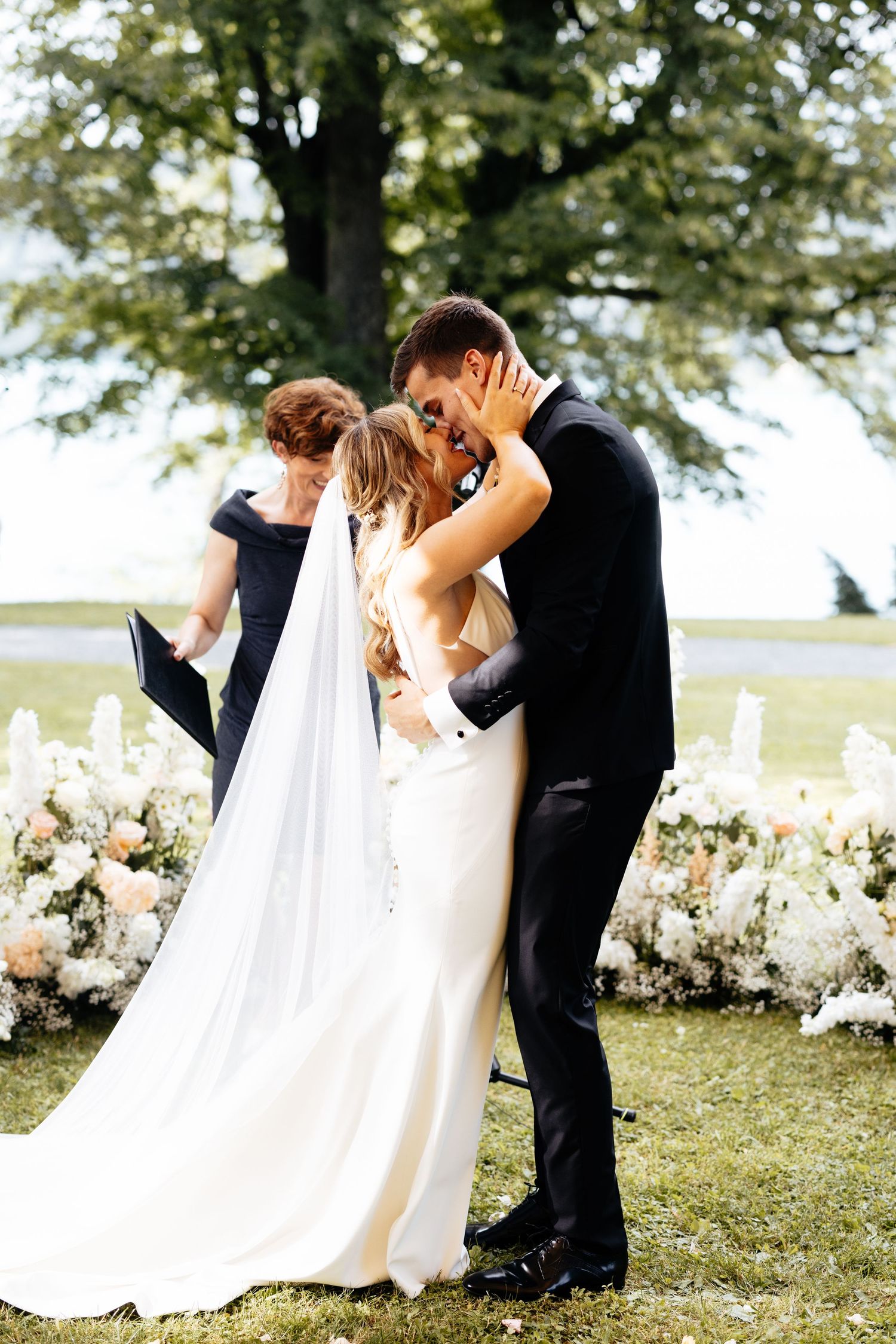 A romantic wedding couple shares an intimate kiss in a garden setting surrounded by elegant white floral arrangements.