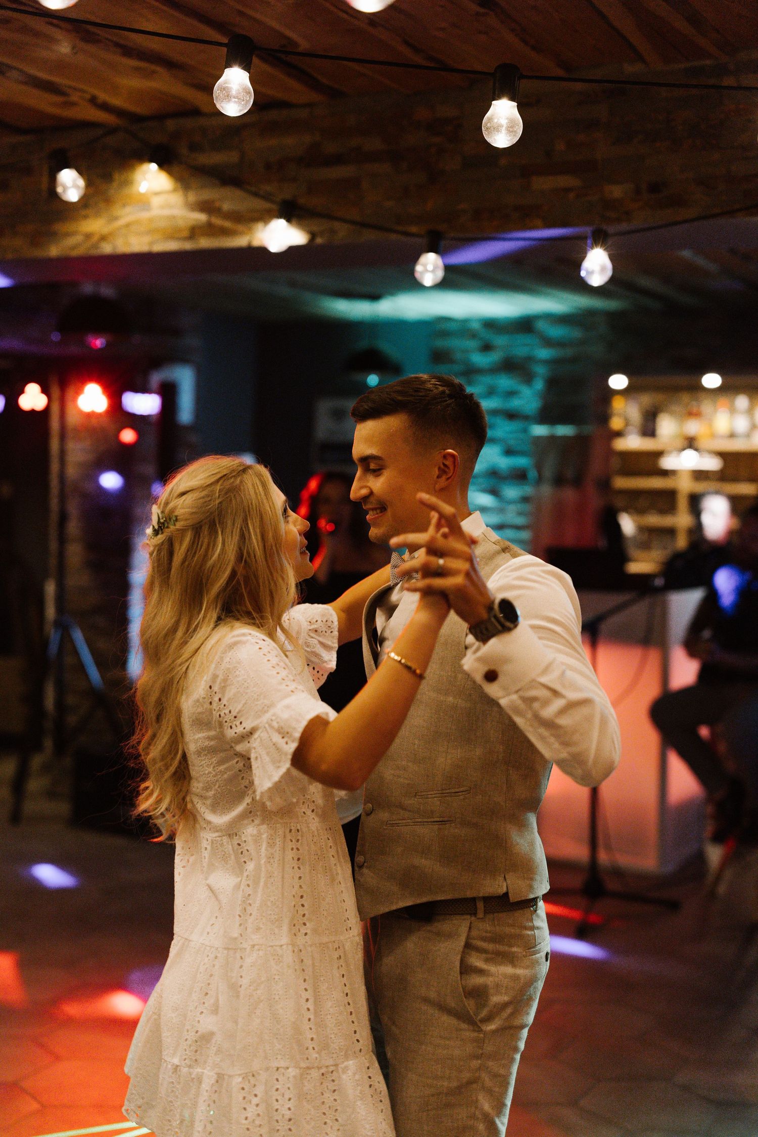 Couple dancing together in a dimly lit venue with string lights and colorful ambient lighting.