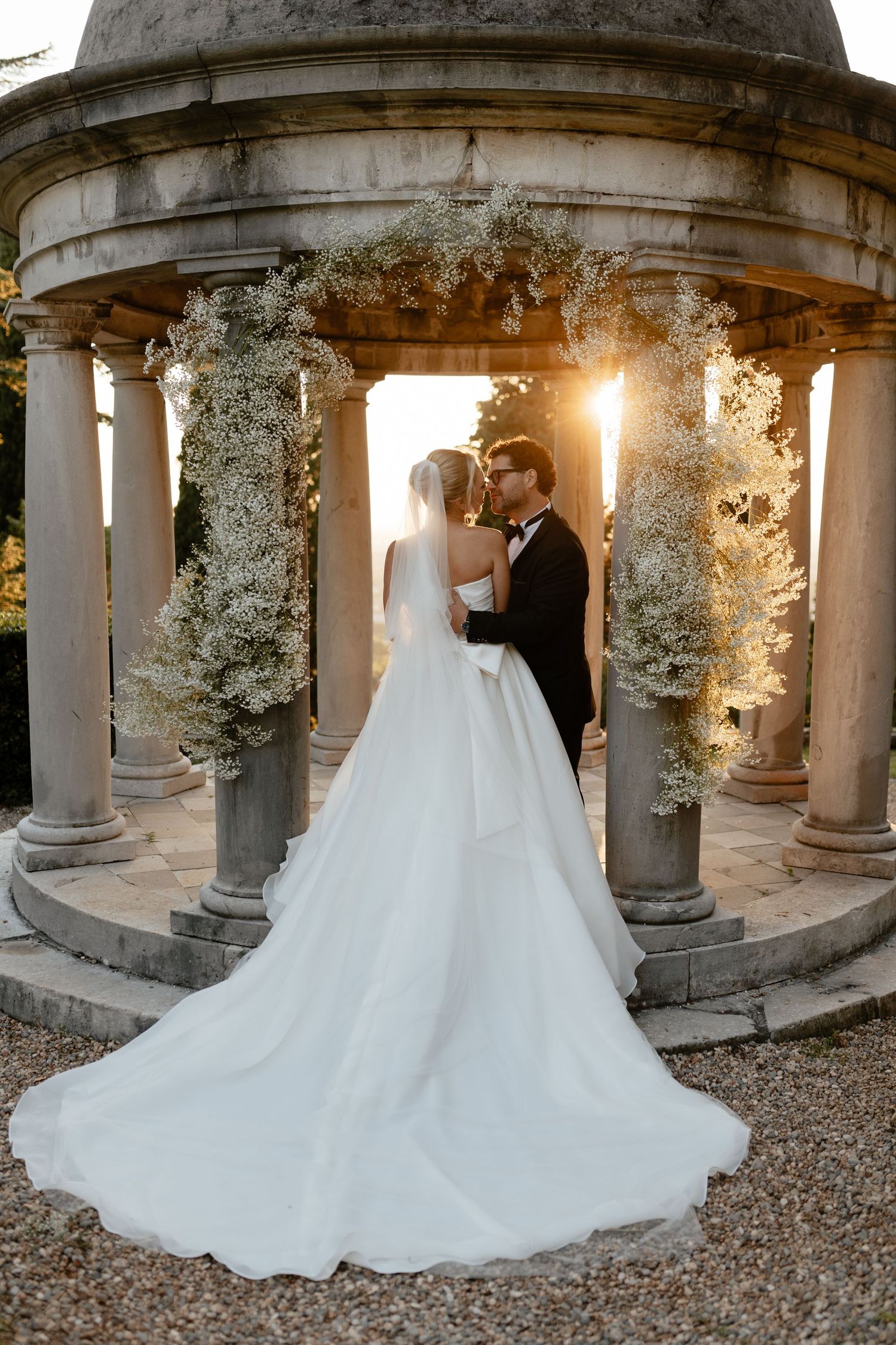 A romantic wedding couple stands in a stone colonnade surrounded by white floral arrangements and gravel ground at sunset.