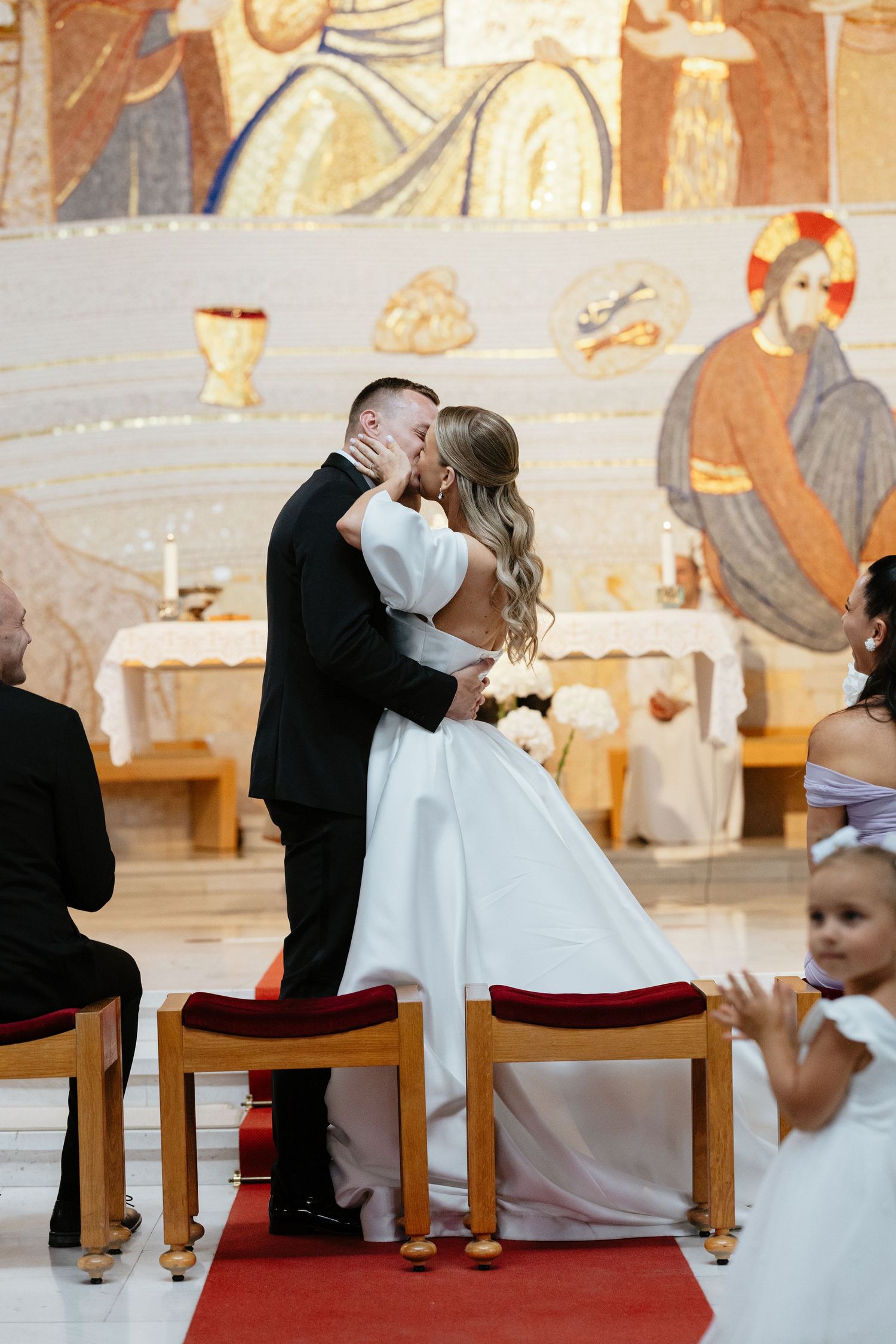 Romantic kiss during wedding ceremony with religious mural in background.