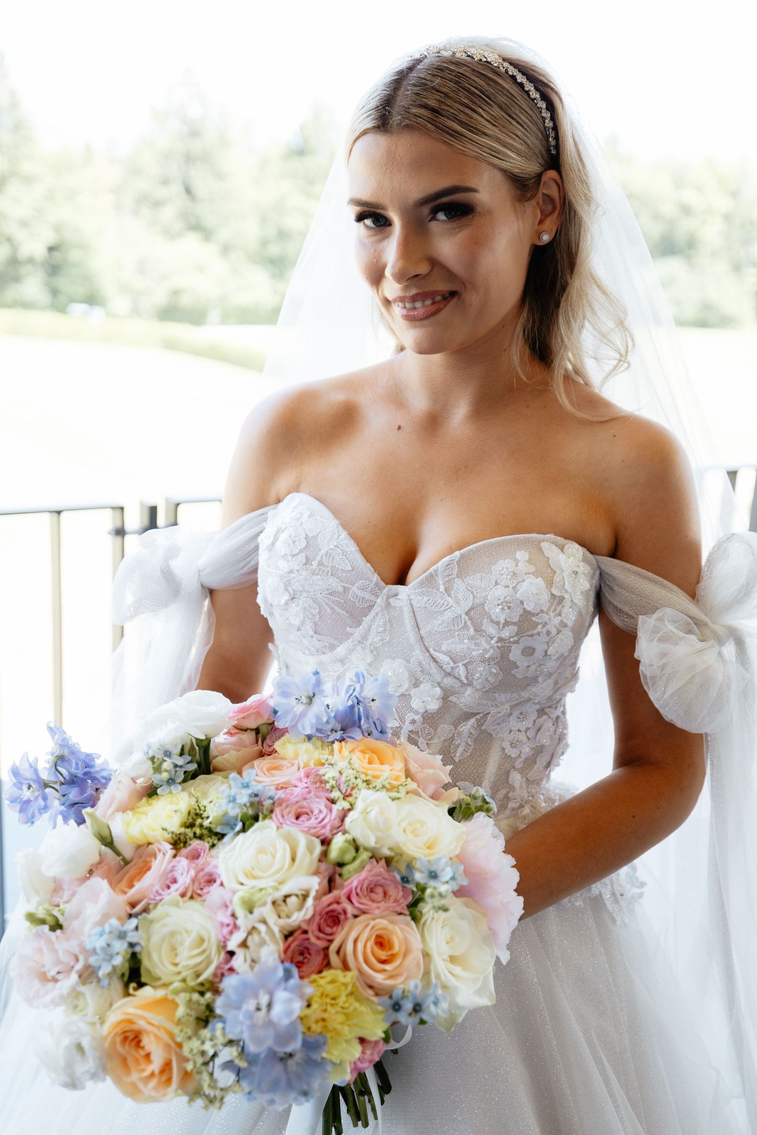 Bride in strapless lace gown holds pastel bouquet of roses and hydrangeas on outdoor balcony.