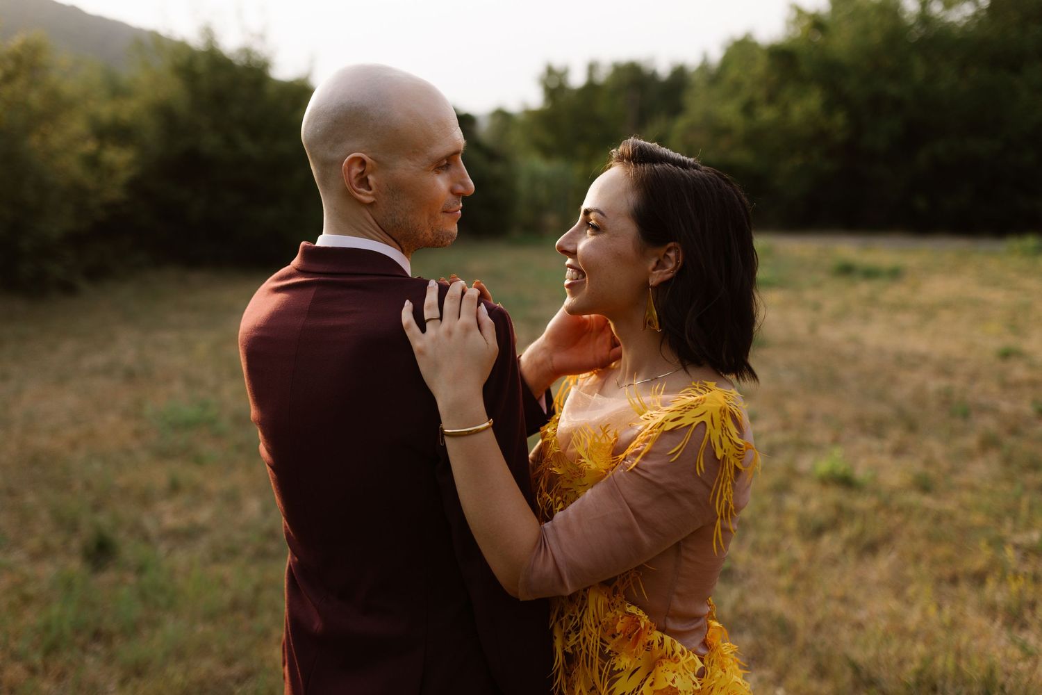 Couple in romantic embrace stands together in golden sunlit field during sunset.