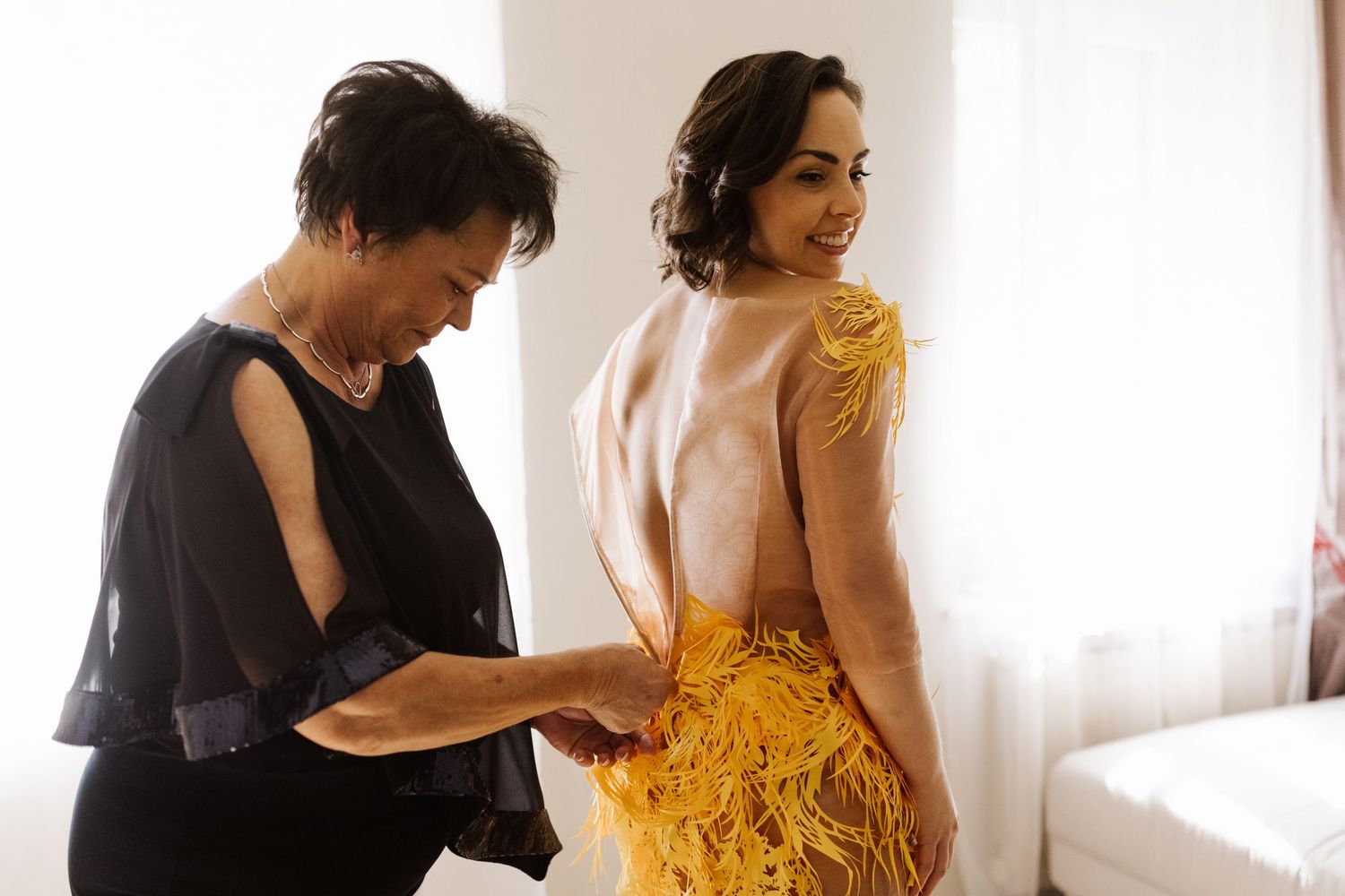 A woman in a stunning yellow feathered gown gets help with final adjustments in a sunlit room.