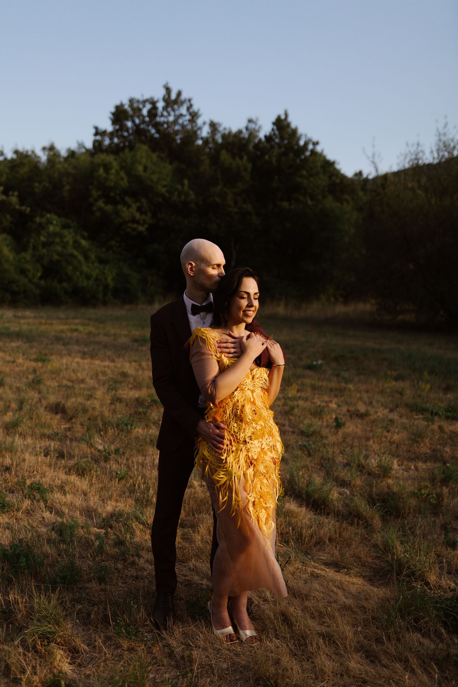 Couple in formal wear with yellow dress posing in a sunlit meadow during golden hour.