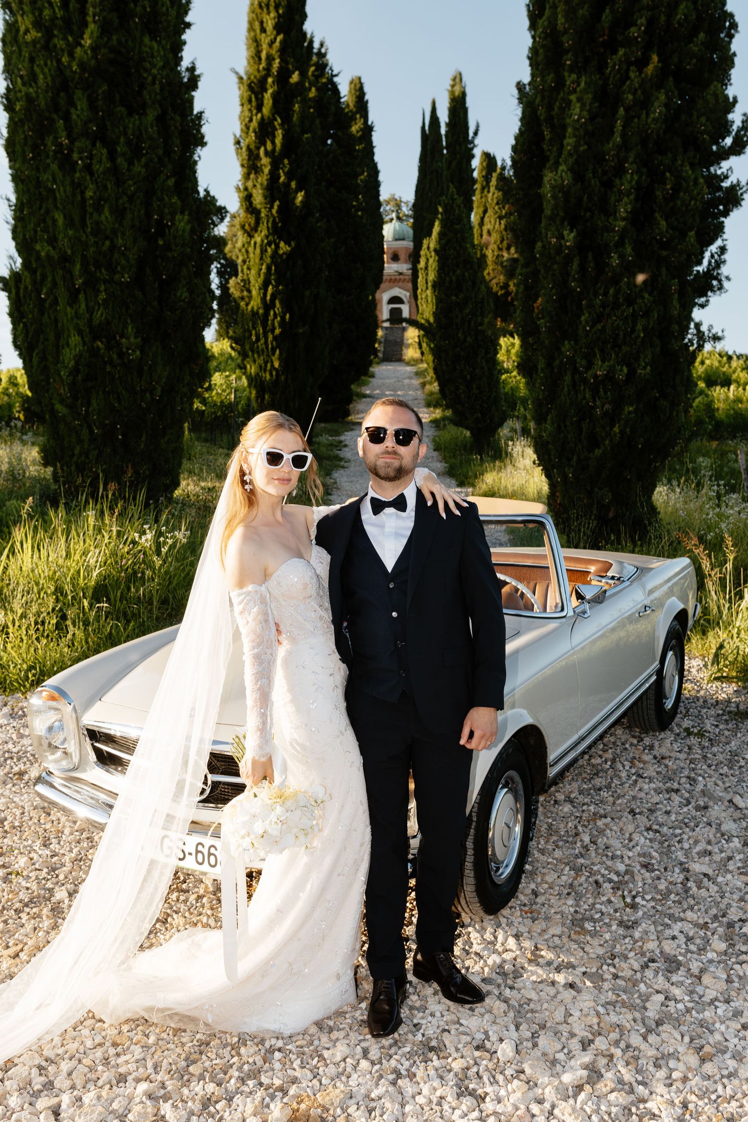 Stylish newlyweds pose with a vintage convertible car among cypress trees in Tuscany.