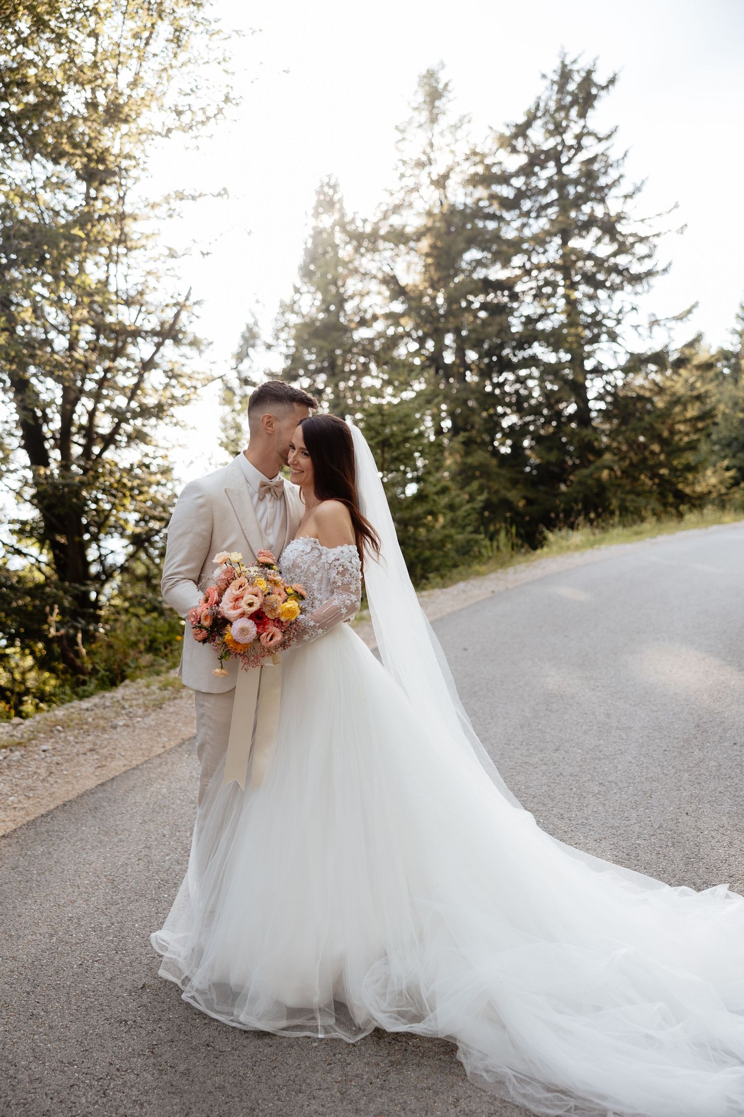 Romantic wedding couple embracing on a tree-lined mountain road with a flowing white dress and autumn bouquet.