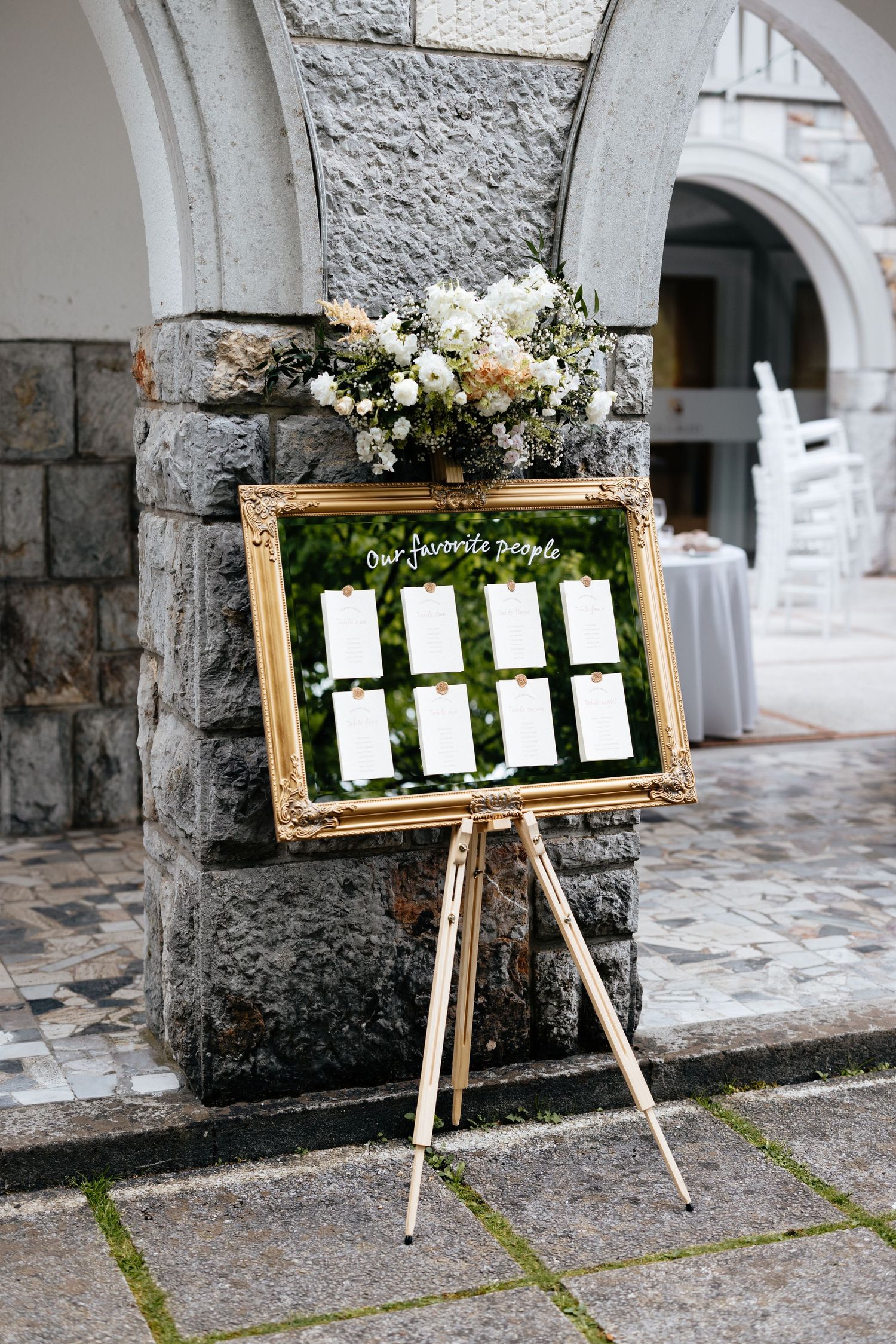 An ornate gold-framed seating chart display on an easel against a stone wall at a wedding venue.