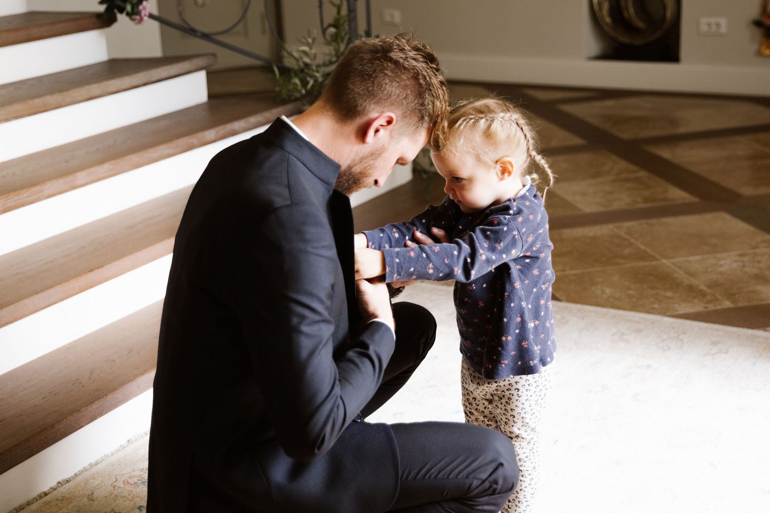 A parent in a dark suit kneels down to share a tender moment with a small child wearing a blue dress.