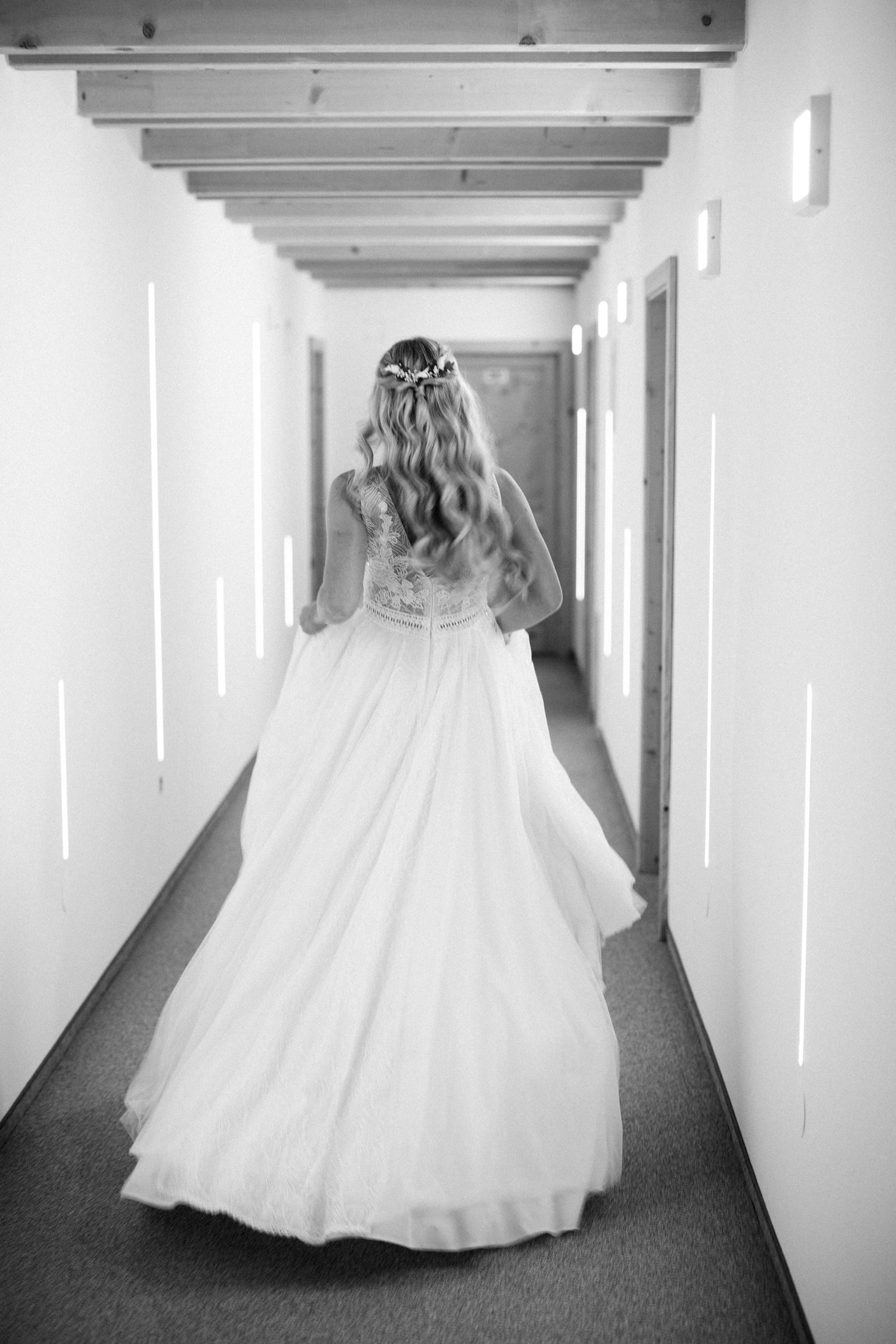 A bride in a flowing white wedding gown stands in a minimalist hallway with modern lighting.