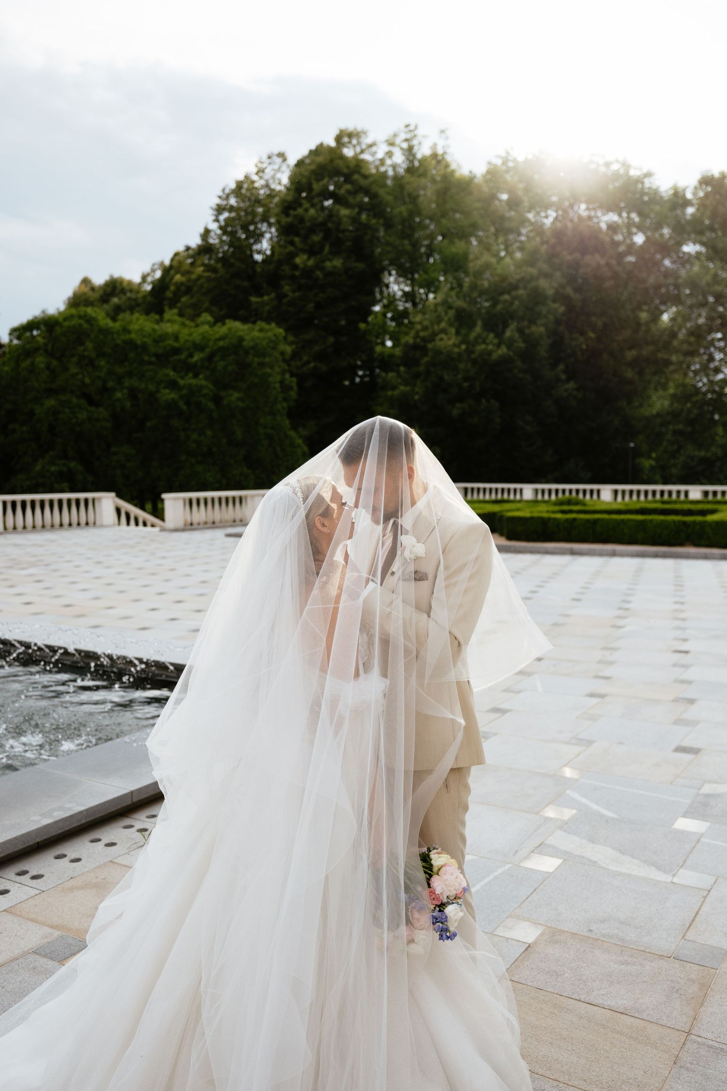 A romantic moment under a veil near a fountain during sunset at an elegant venue.