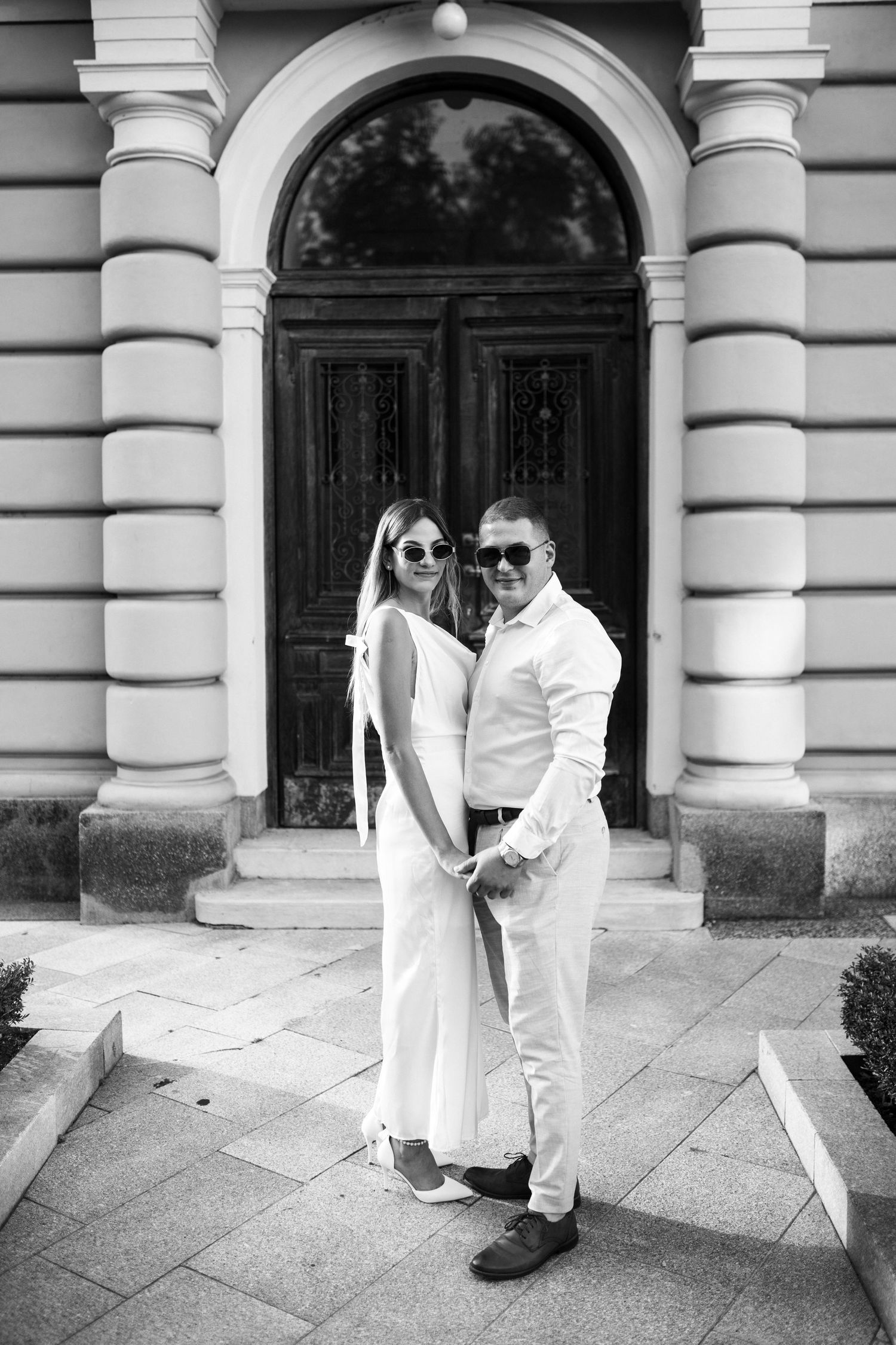 A stylish couple in all-white outfits posing under an ornate archway entrance.