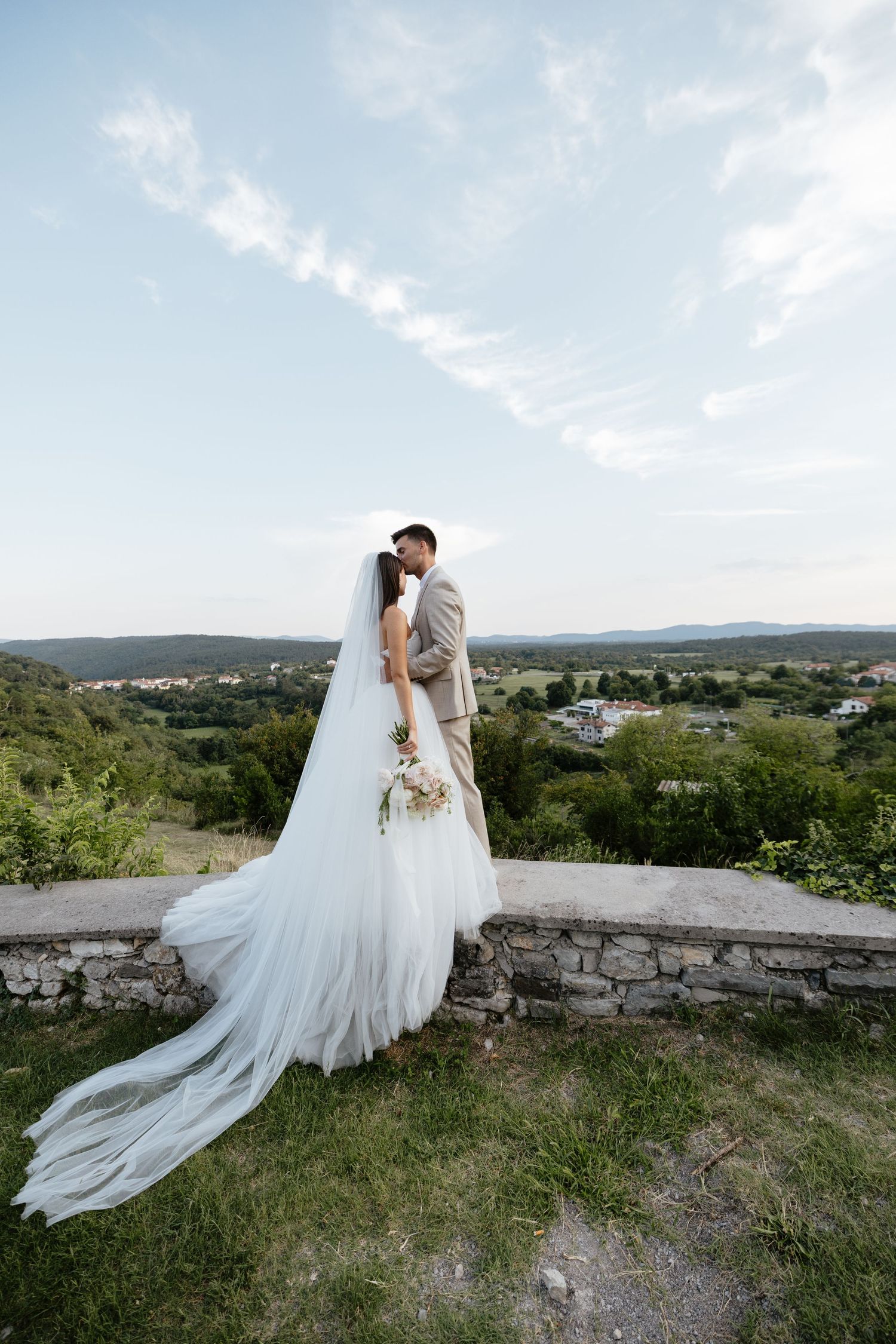 Wedding couple embraces on stone wall overlook with flowing veil and scenic countryside view.