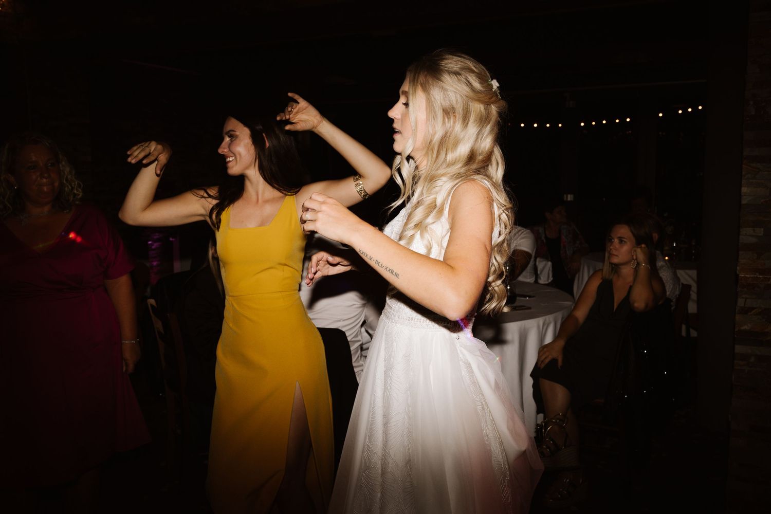 Wedding party dancing and celebrating in a dimly lit reception venue with string lights overhead.