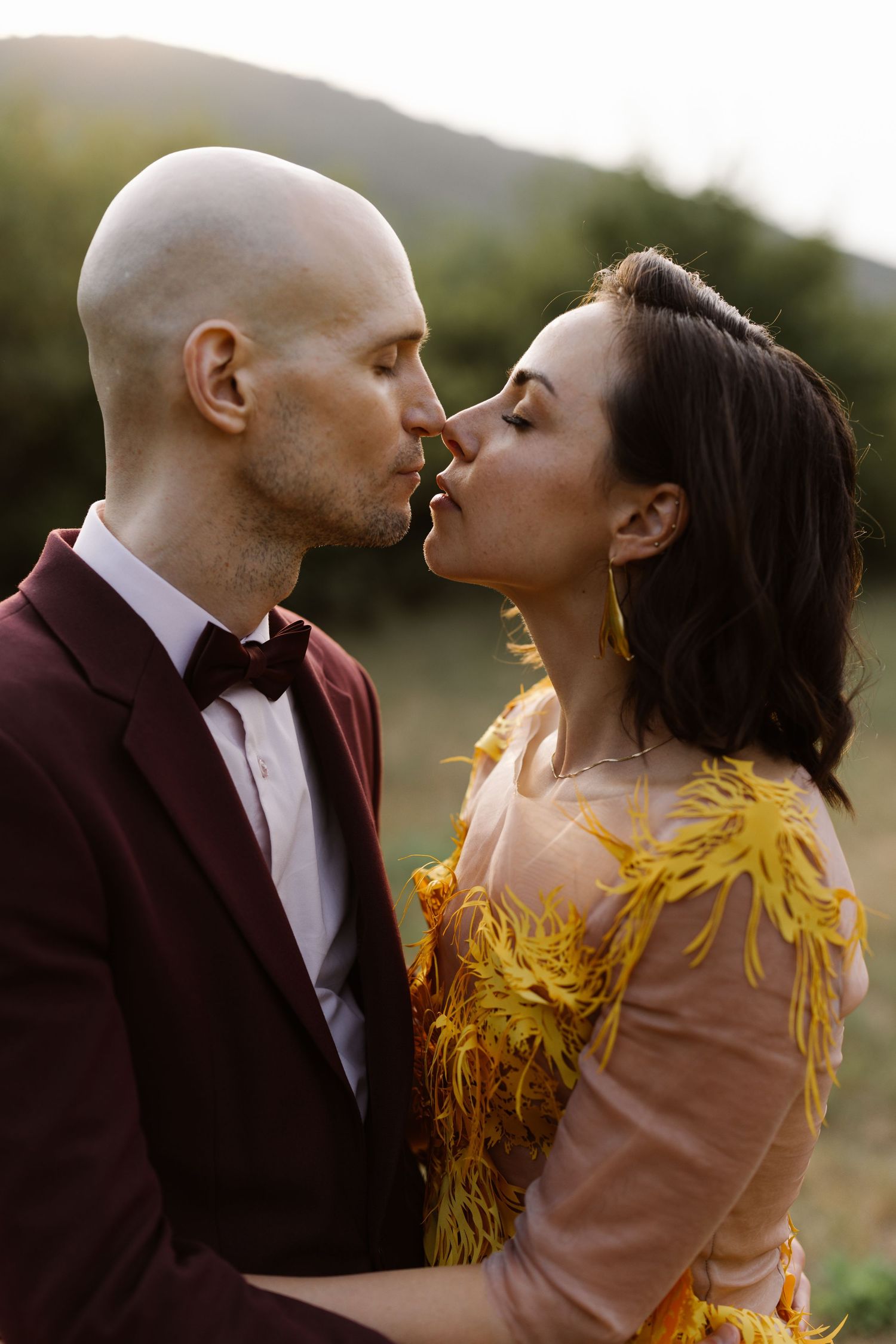An intimate moment between a couple wearing burgundy and yellow formal attire against a natural outdoor backdrop.