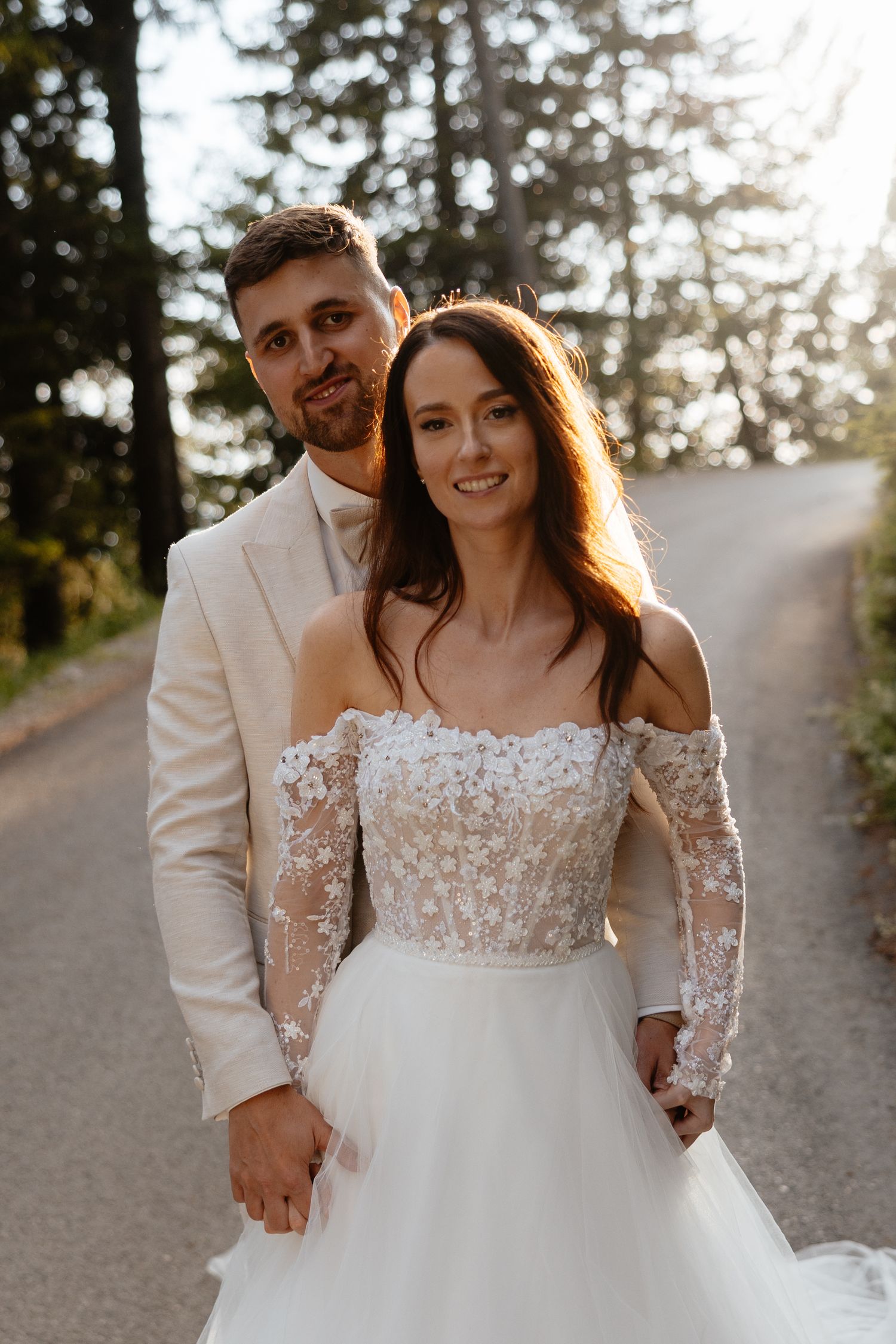 A couple walks hand in hand on a sunlit path wearing elegant wedding attire.