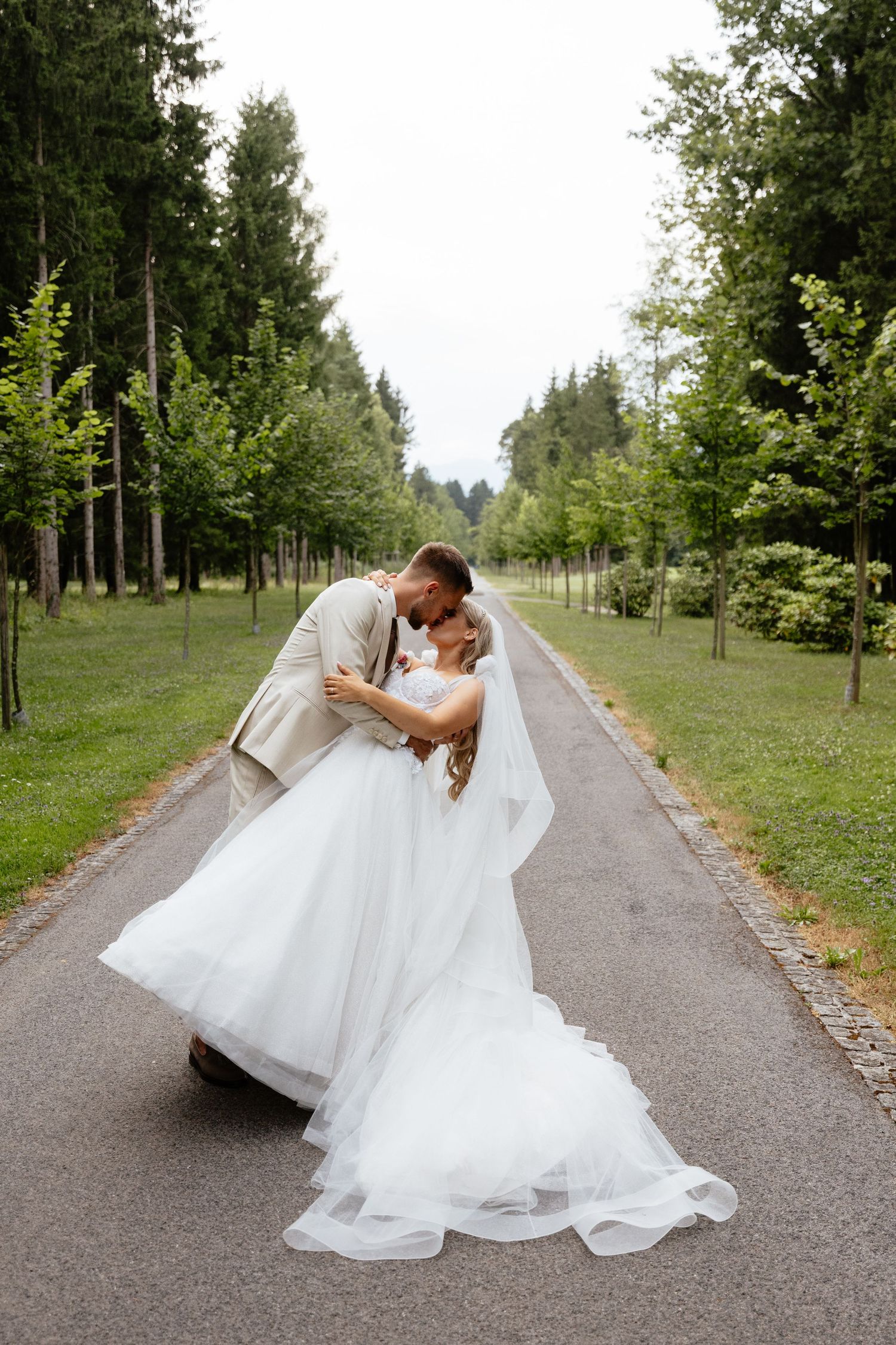 Romantic wedding couple dips and poses on tree-lined path in outdoor setting.