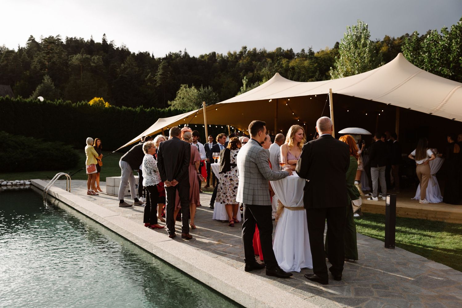 Outdoor wedding reception under beige canopy tents with guests gathered near a lake.