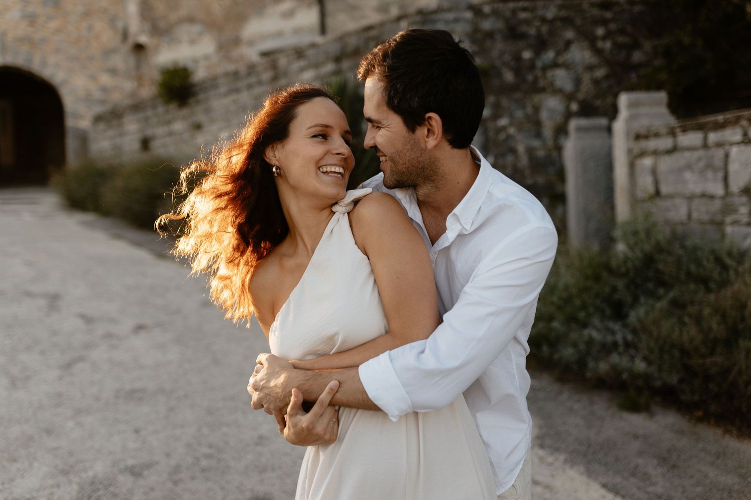 A couple embraces and laughs together while walking along a stone wall during a romantic outdoor photoshoot.