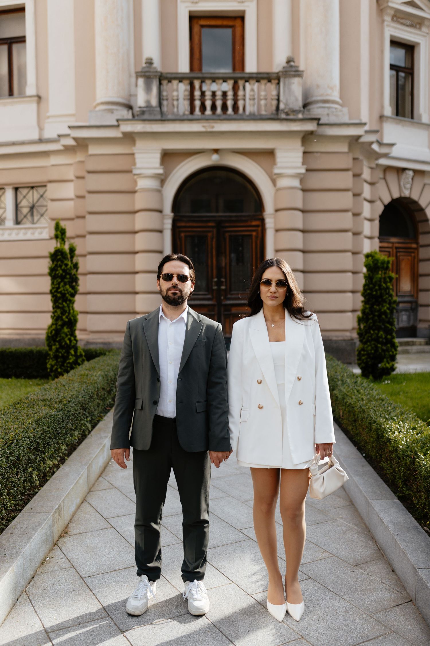 A stylish couple in formal attire poses in front of a classic building entrance.