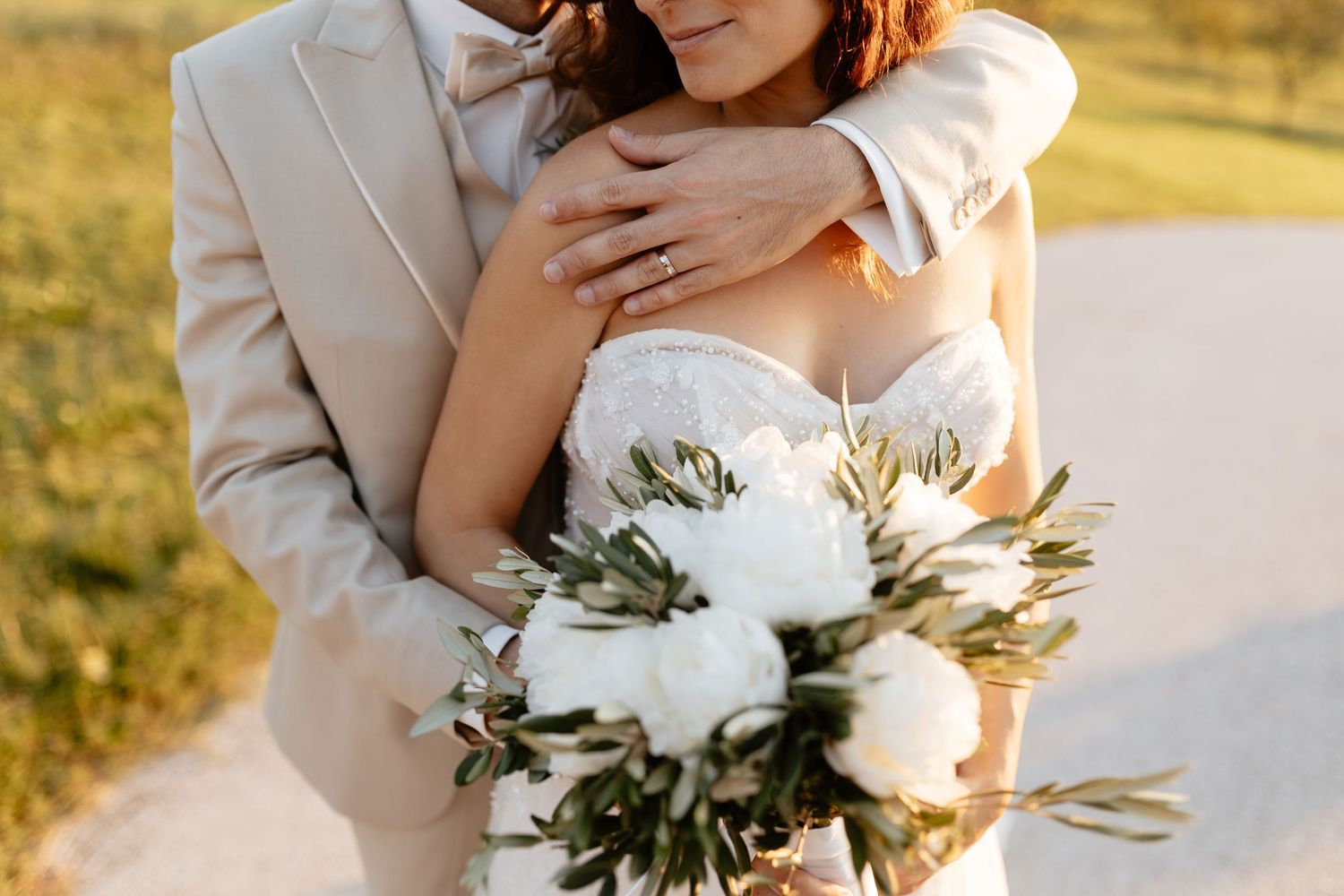 A romantic wedding couple embraces in golden sunlight while holding a white peony and greenery bouquet.