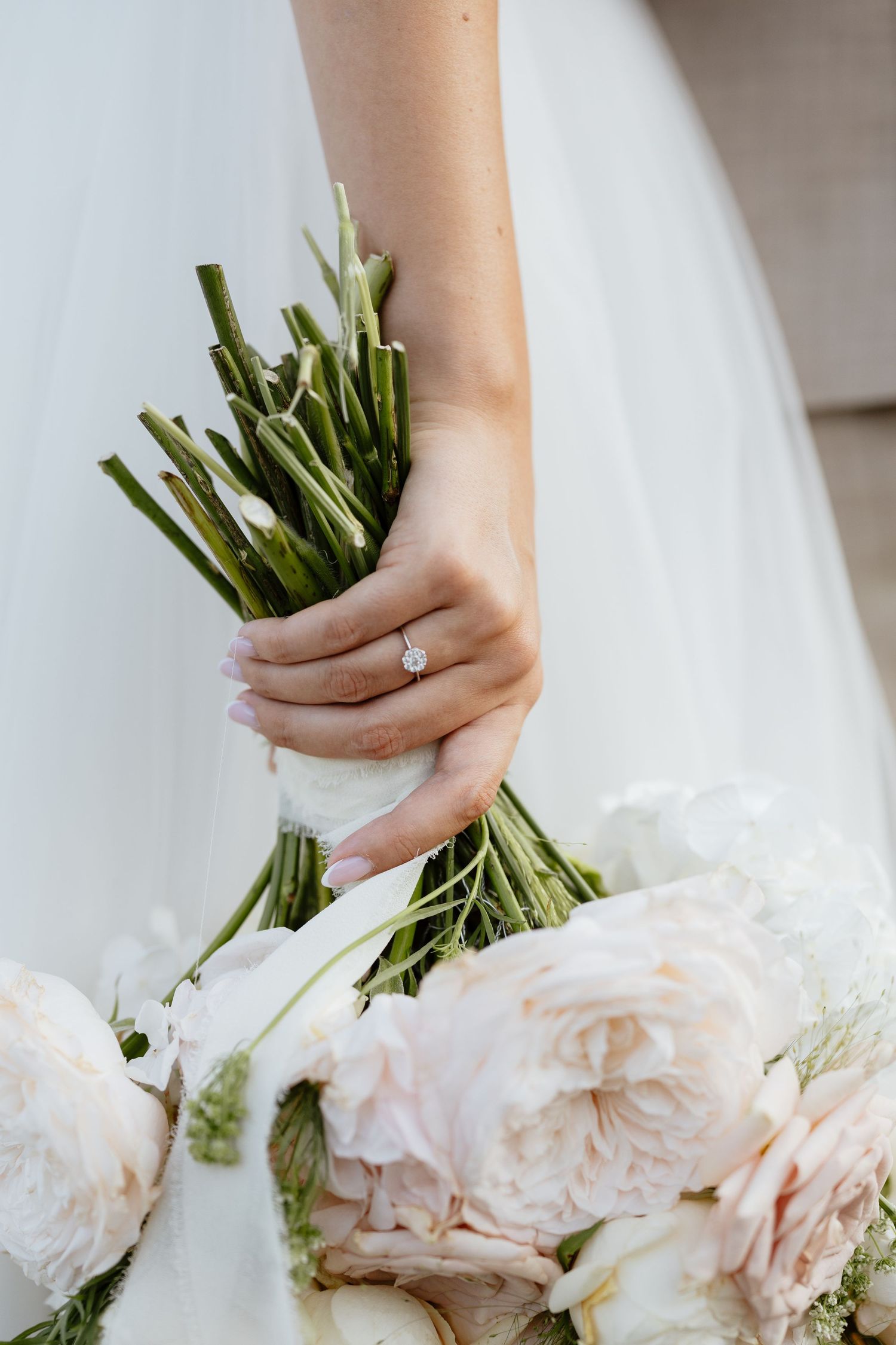 Close-up of hands holding elegant bouquet with blush roses and white flowers against white dress.