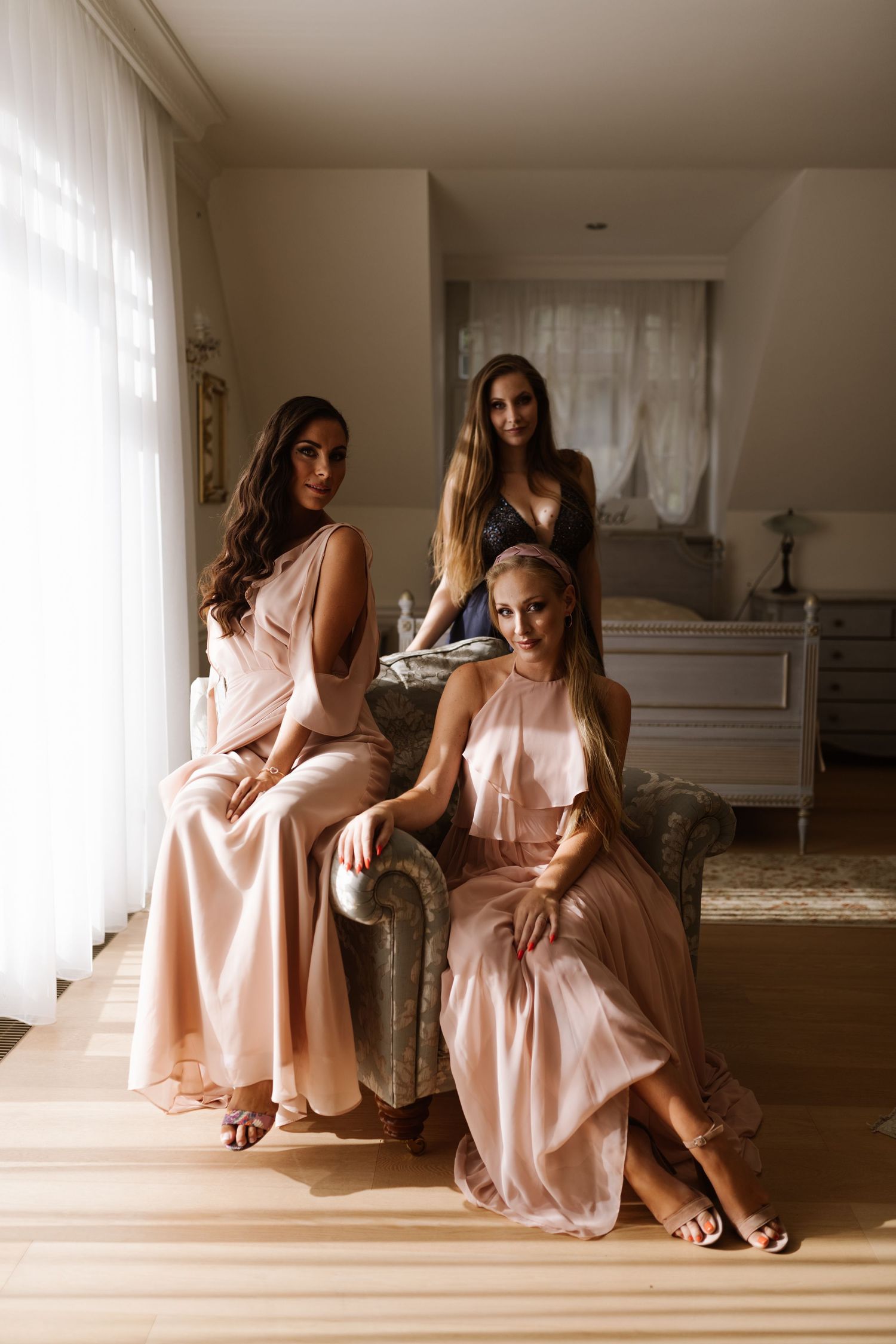 Three bridesmaids in blush pink dresses pose elegantly in a sunlit room with a white piano.