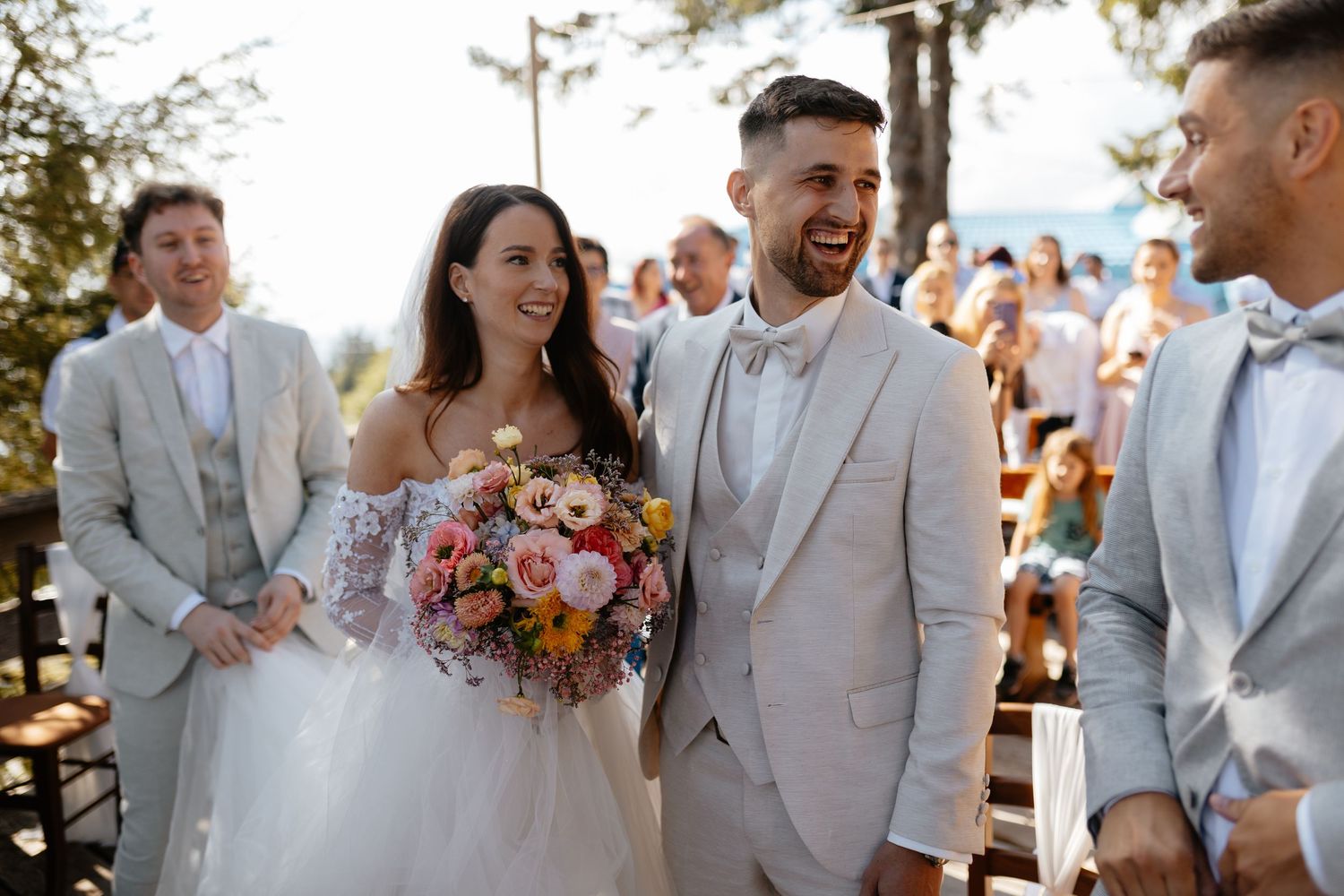 A joyful outdoor wedding ceremony with guests gathered under trees as the couple shares a happy moment.