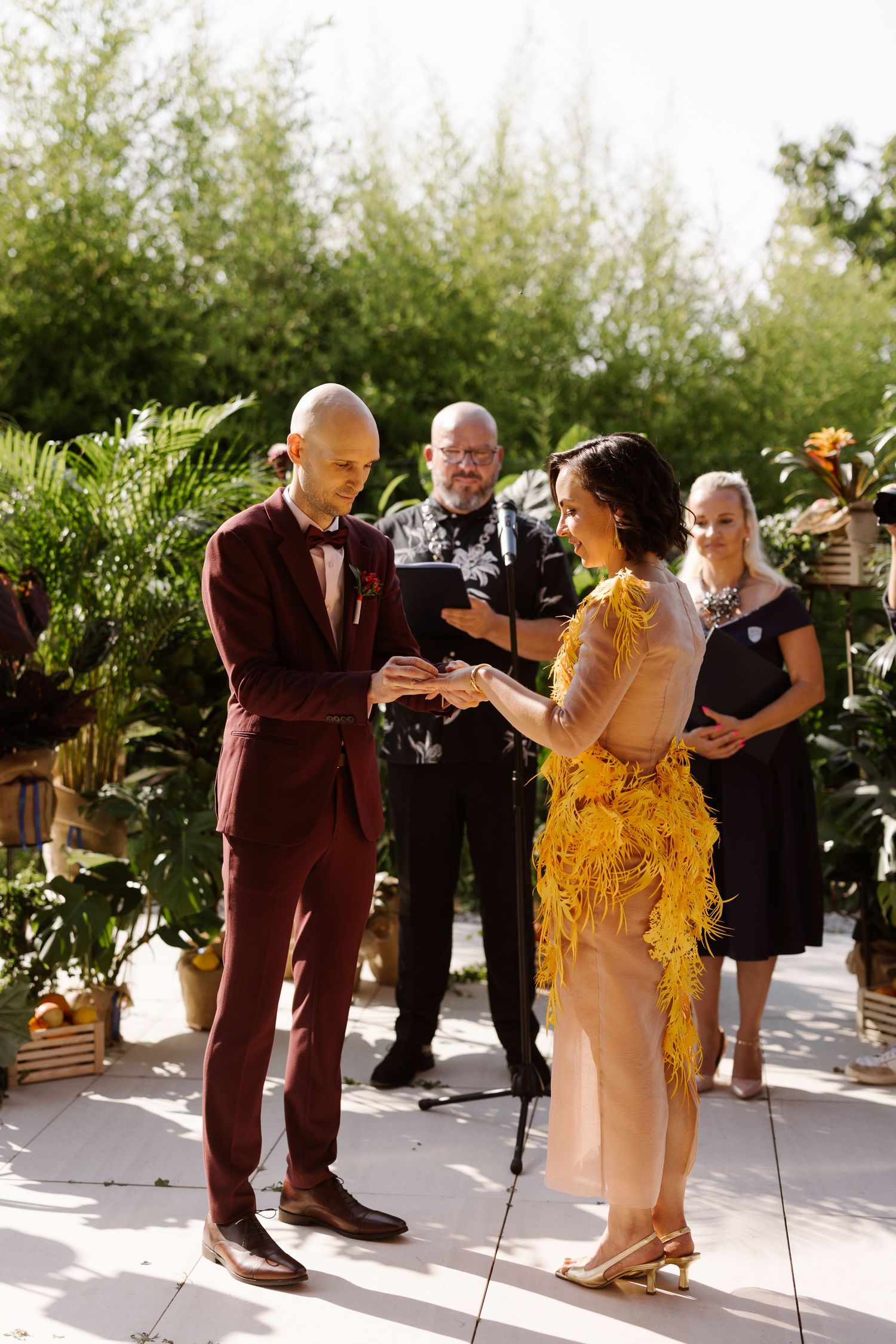 A wedding ceremony takes place outdoors with a couple in burgundy and yellow attire exchanging vows in front of an officiant.
