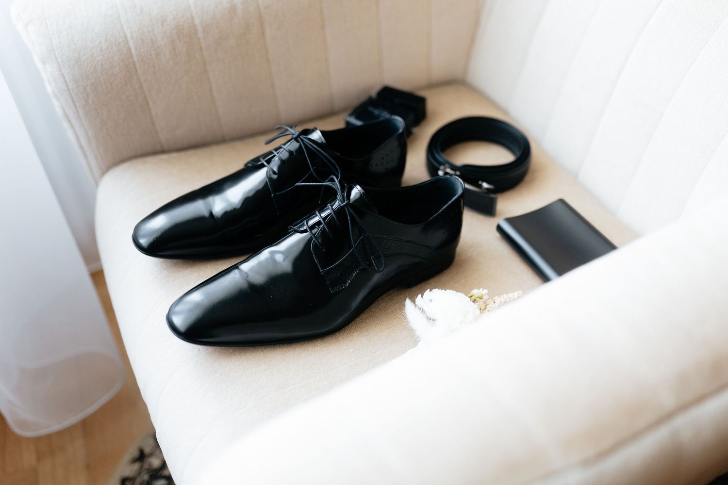 A pair of shiny black leather dress shoes sits on a white surface next to a phone and other items.