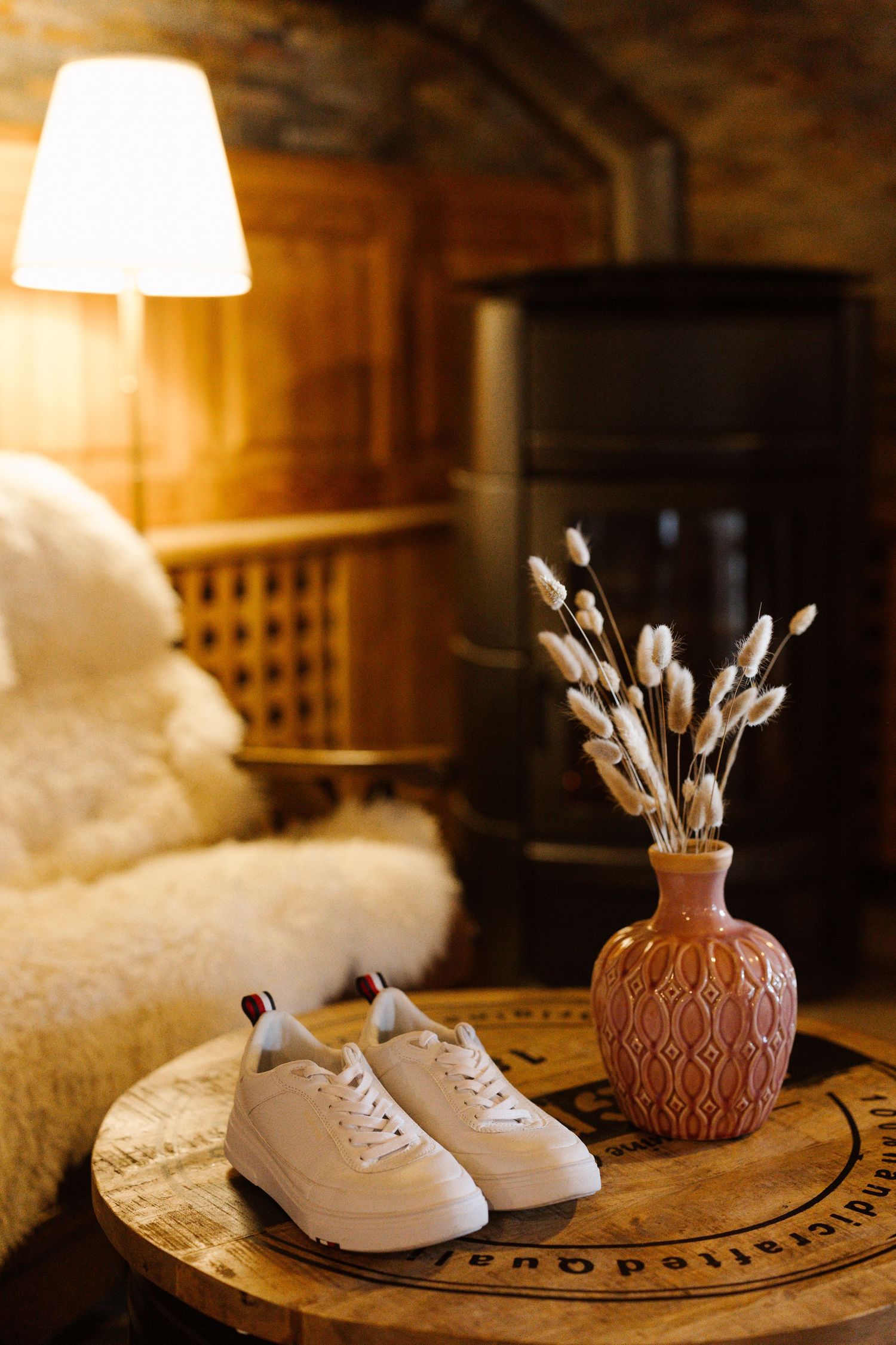 White sneakers and dried pussy willows in a terracotta vase sit on a wooden table with warm lighting.