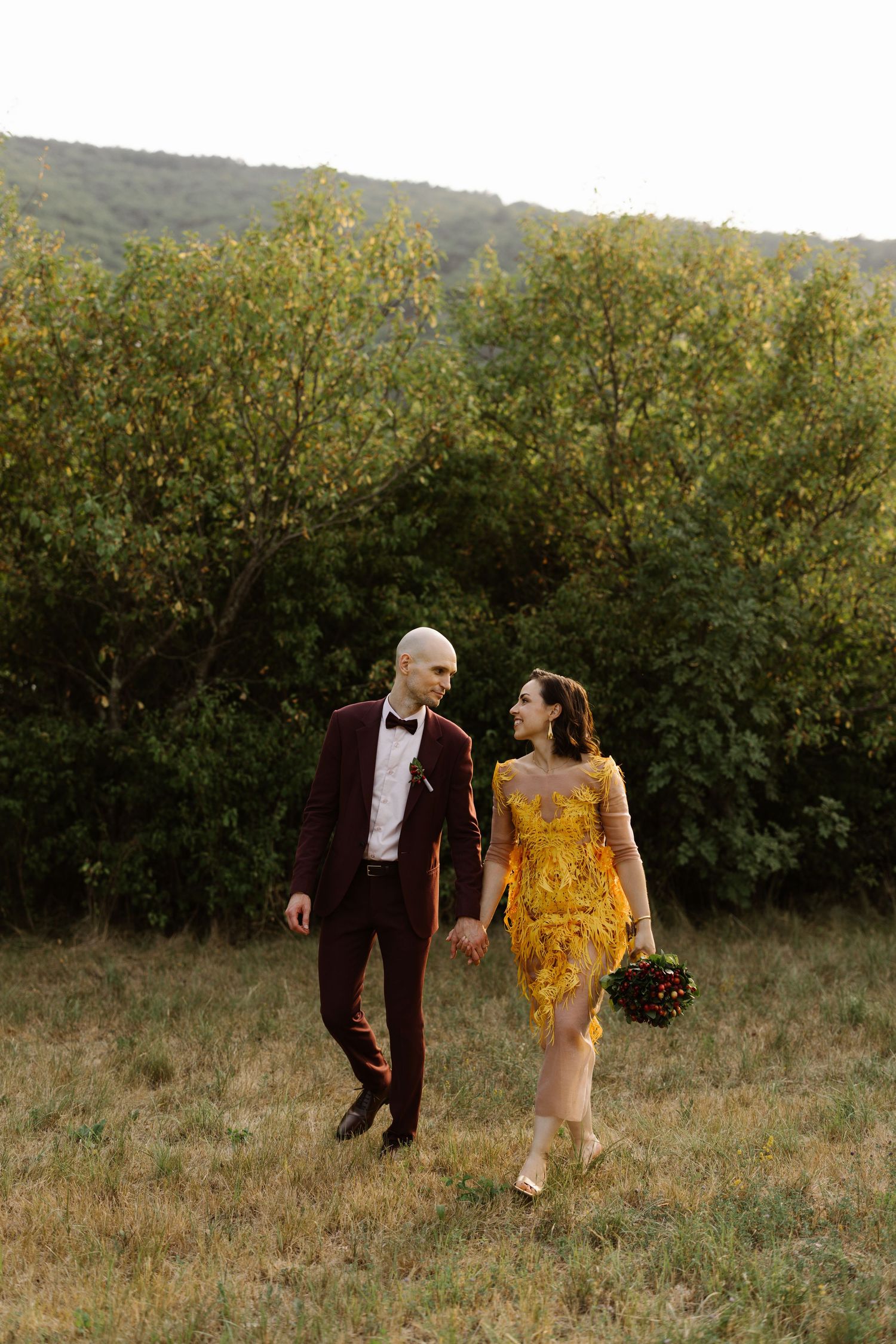 A couple walks hand in hand through a meadow, wearing burgundy and yellow formal attire.