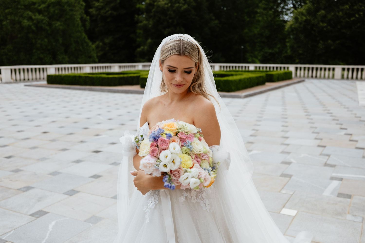Bride holding pastel bouquet poses on ornate white stone patio during wedding photoshoot.